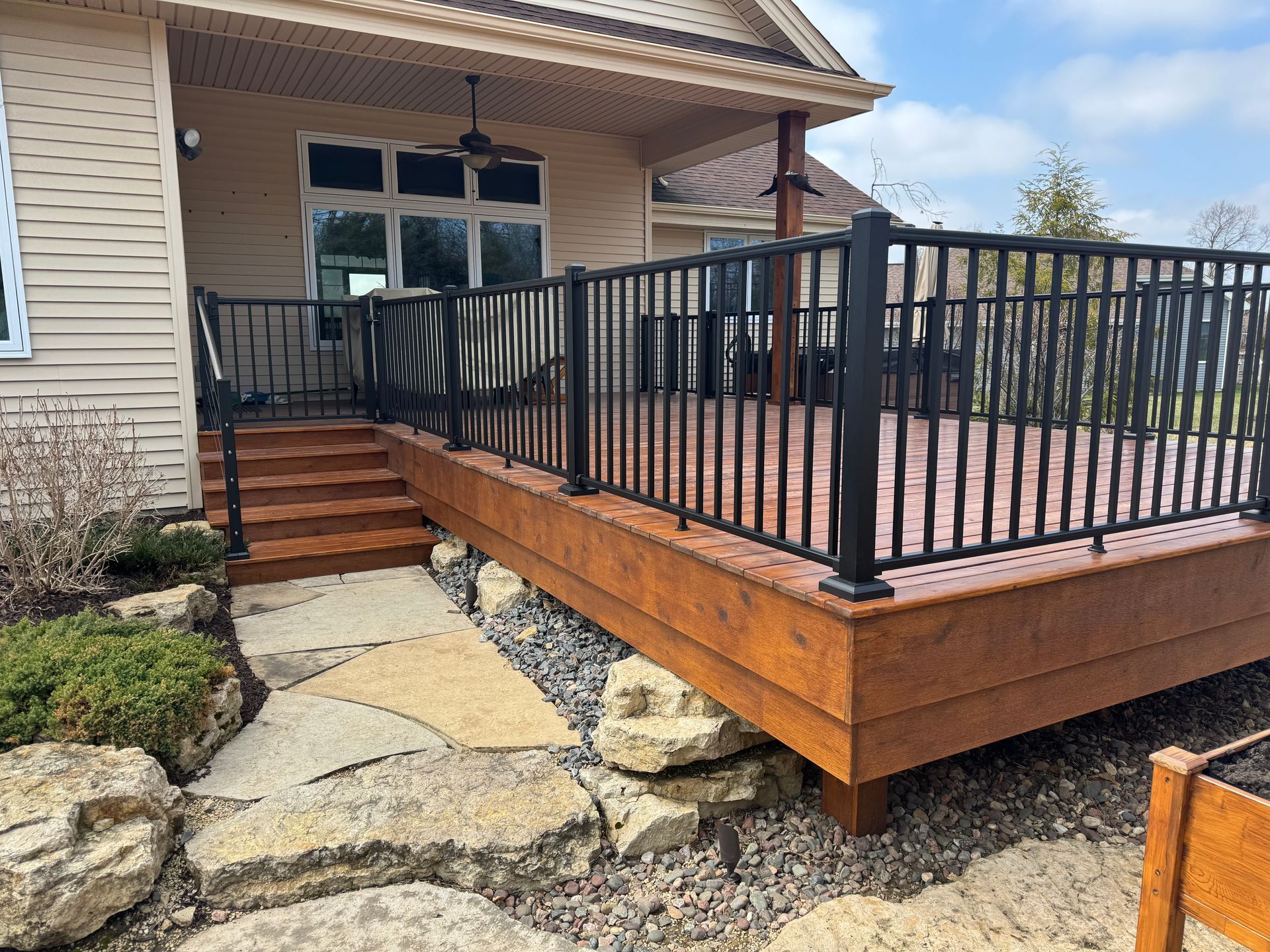 A wooden deck with black railings and steps leading to a house, stone path in the foreground.