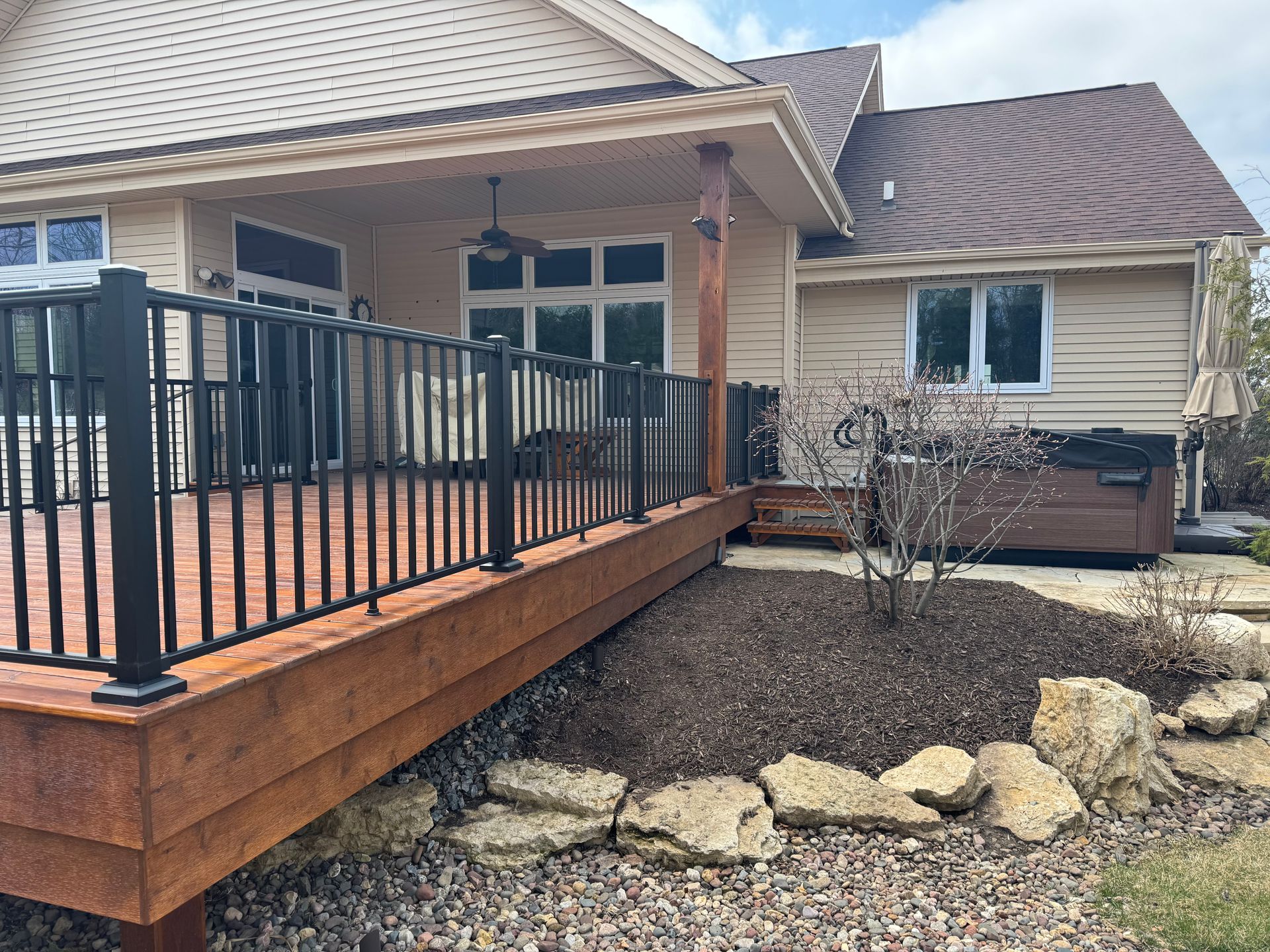 Wooden deck with black railing next to a house with hot tub and landscaping.