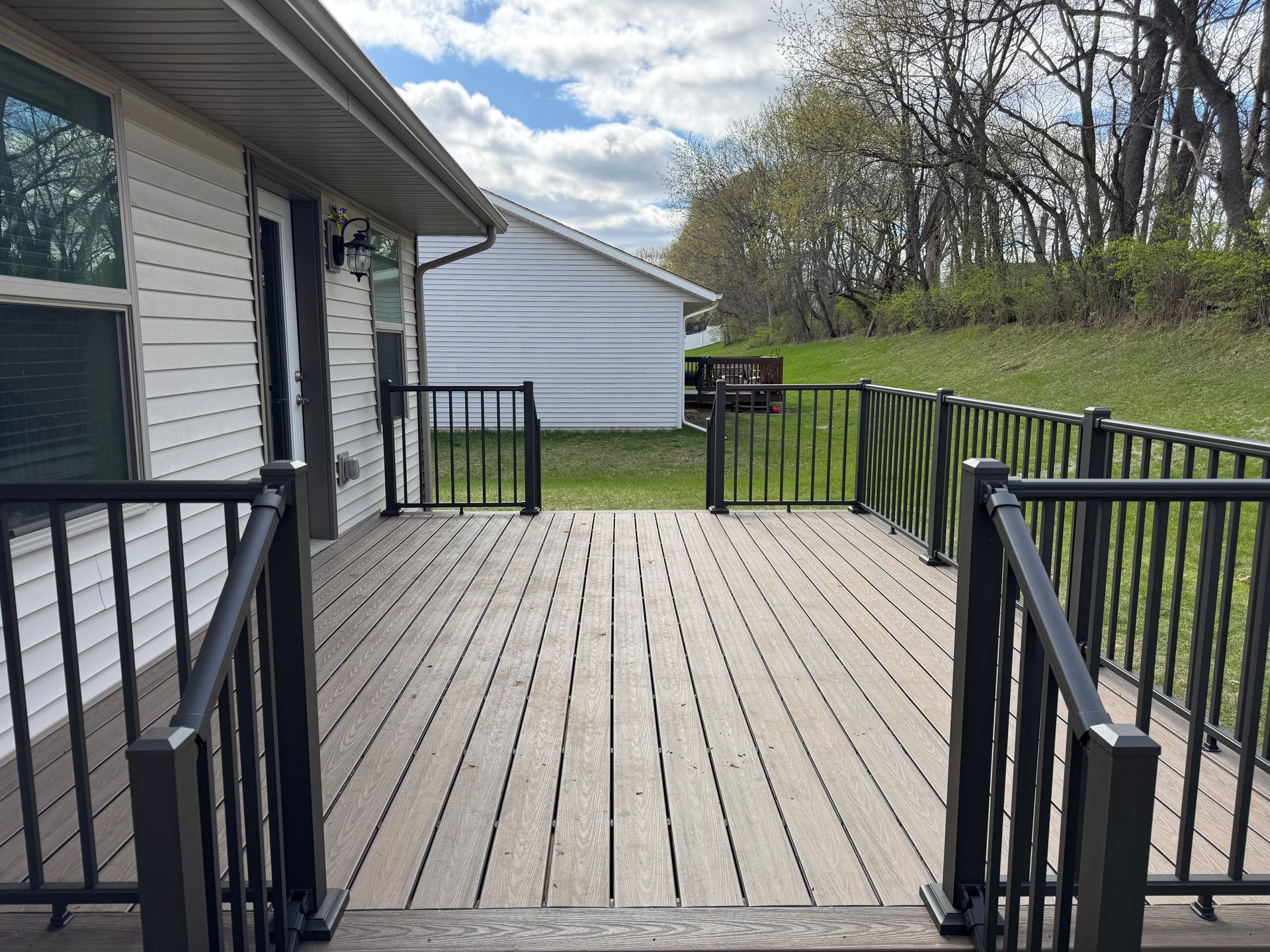 Wooden deck with black railings, leading to a fenced yard; white house in the background.