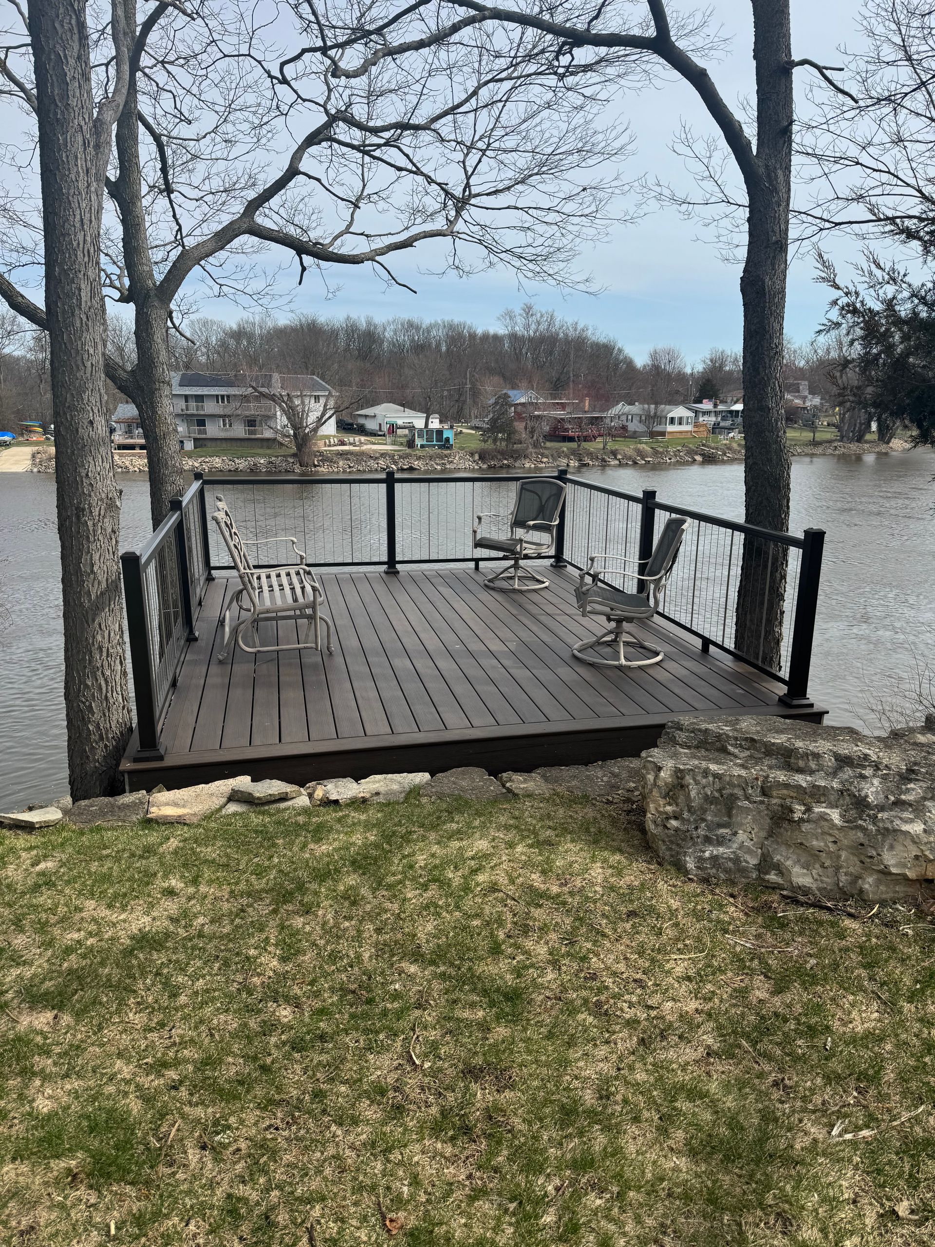 Wooden deck over water with glass railings, small table and chairs, trees and green grass.