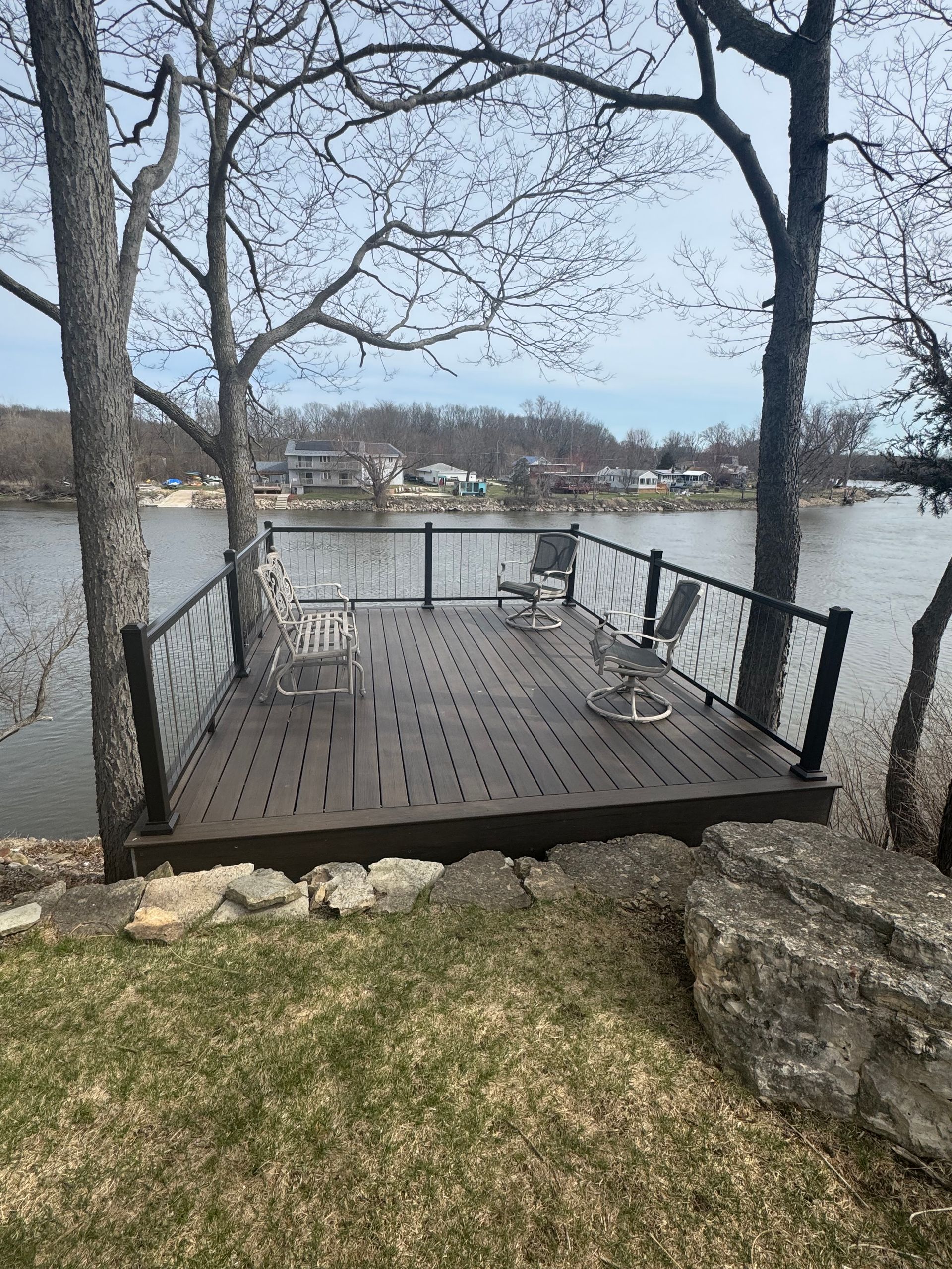 Wooden deck with black railing and chairs overlooking water, trees in the foreground.