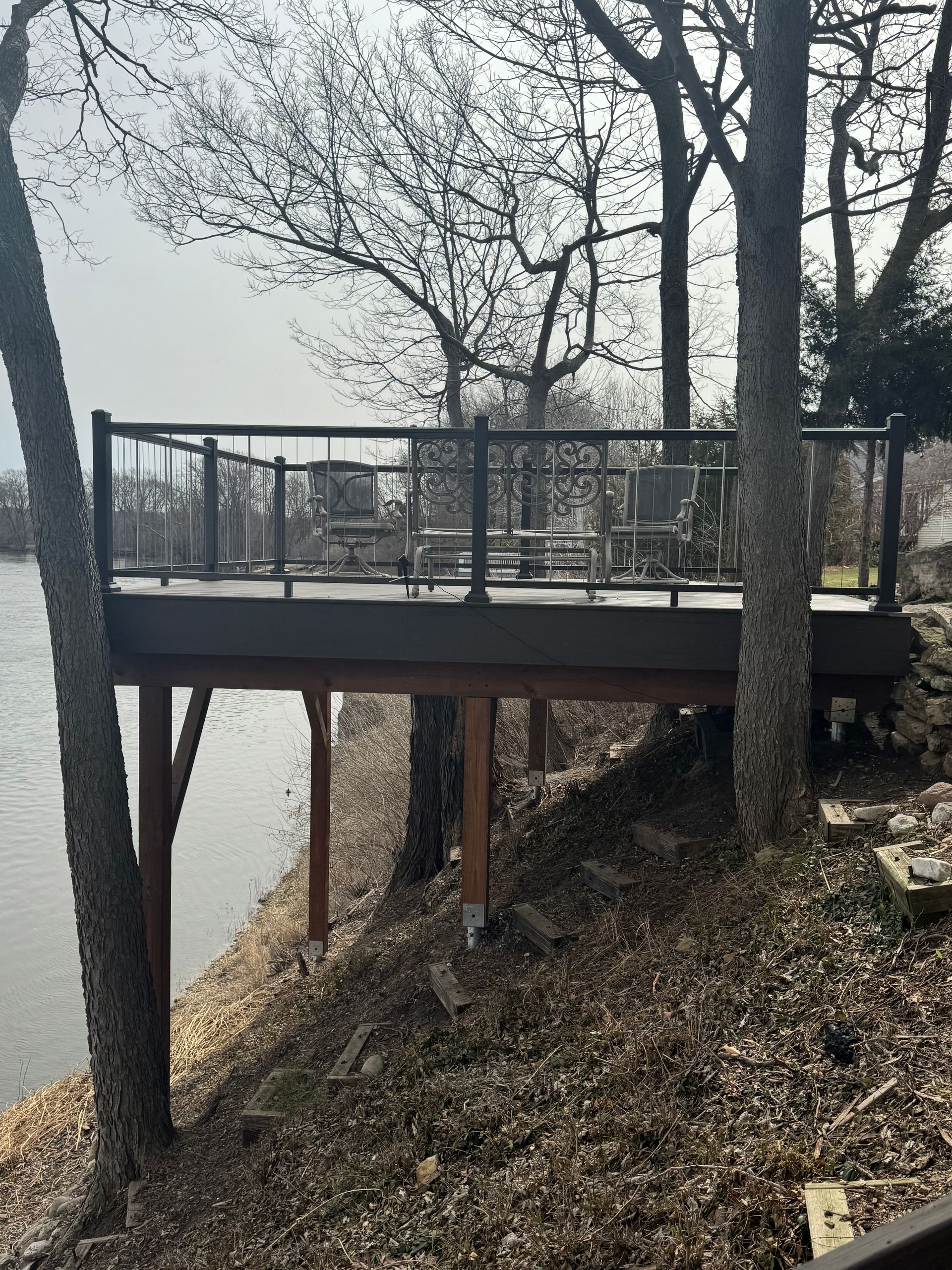 Wooden deck overlooking water, furnished with seating. Supported by posts on a hillside with leafless trees.