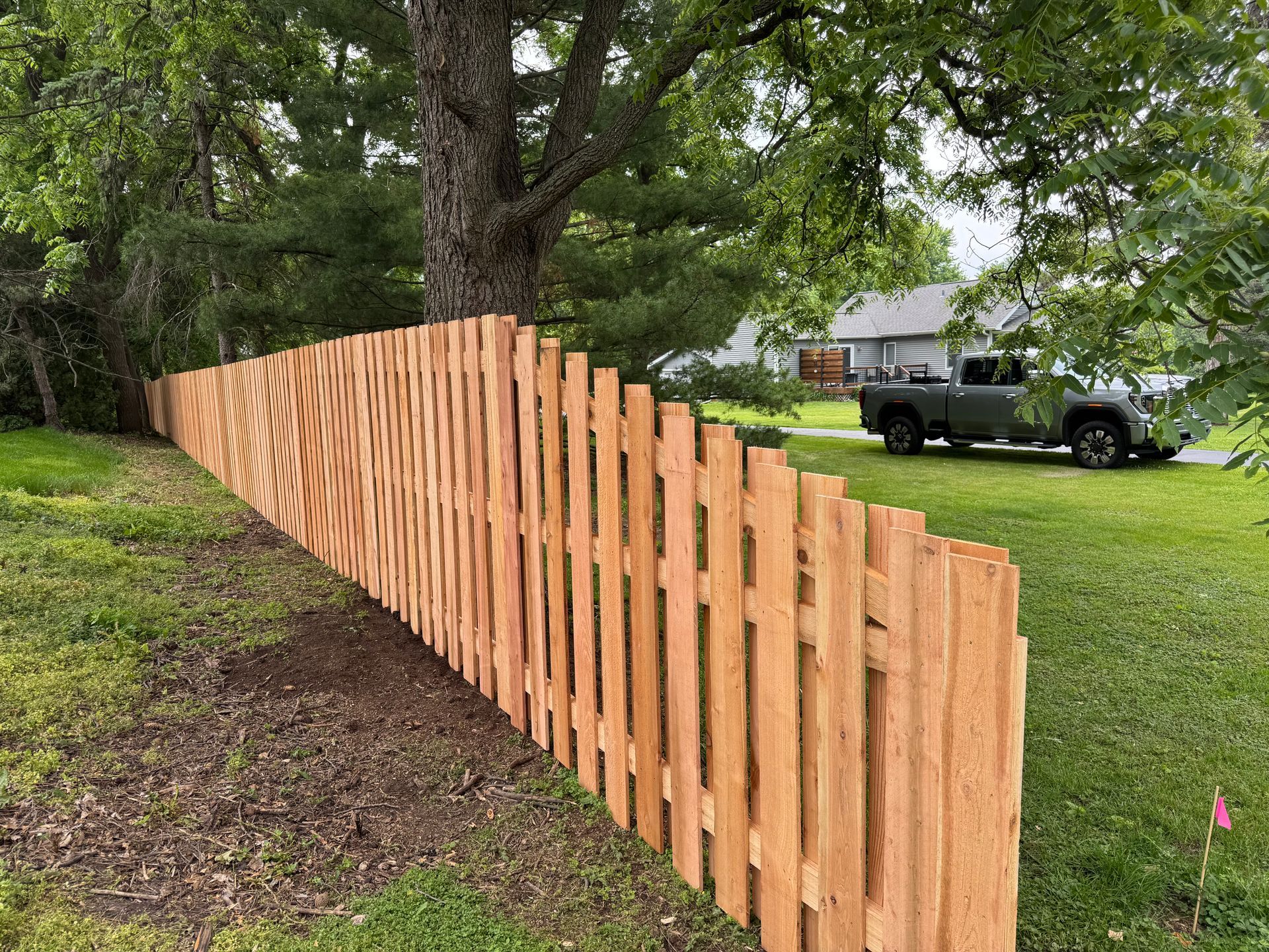 New wooden fence along a grassy yard, under a tree. A gray truck is parked on a lawn in the background.