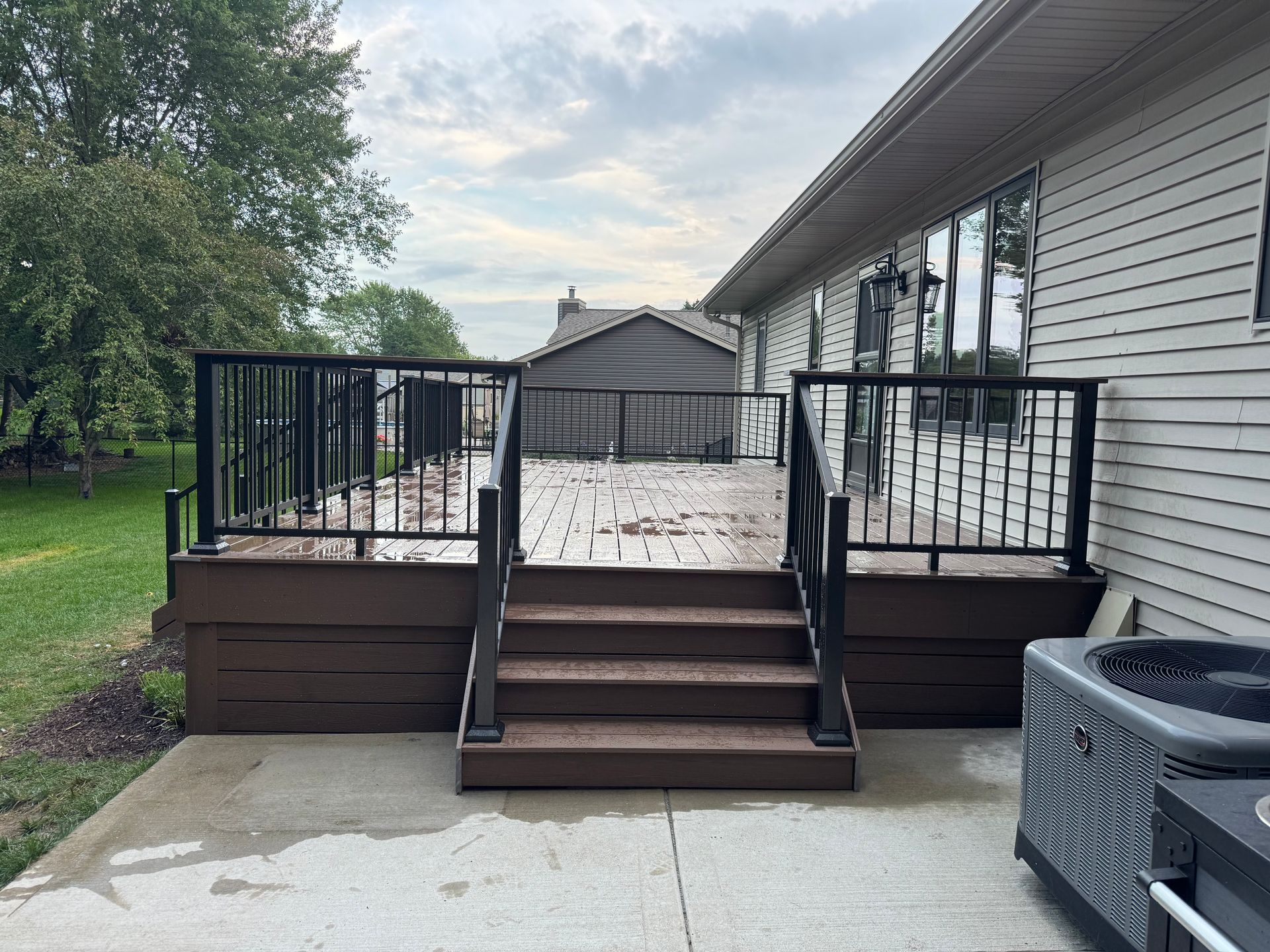 Deck with black railing, brown steps, and composite decking attached to a house with an AC unit.
