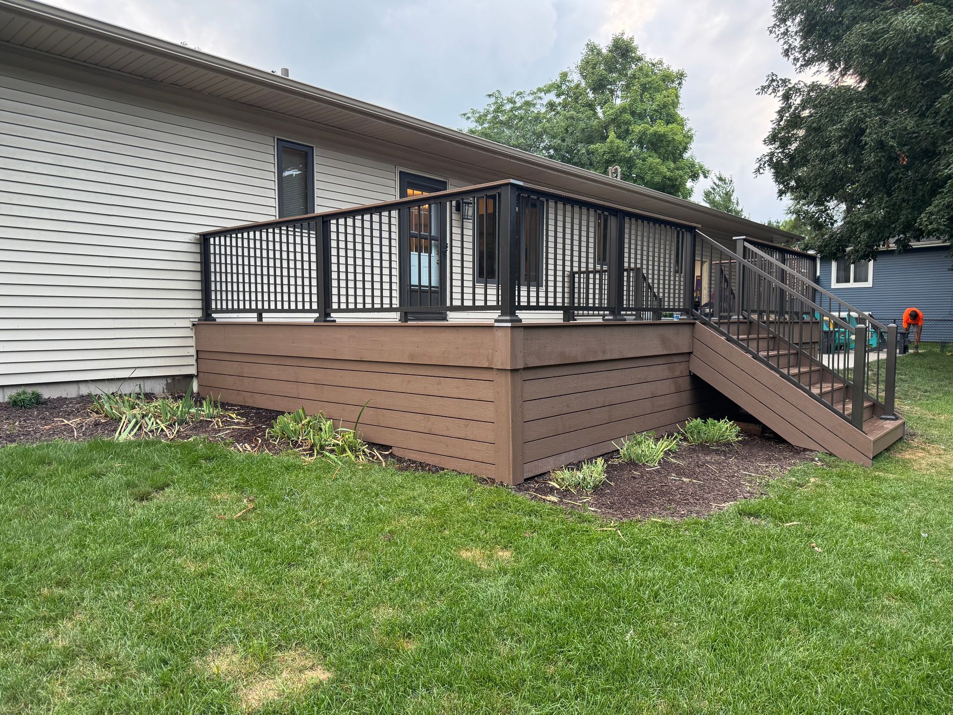 Brown composite deck with black railing attached to a beige house, green grass, and shrubs.