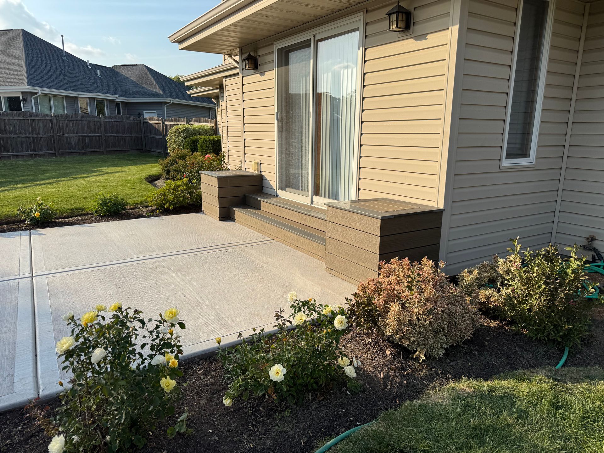 Patio with steps leading to a sliding door; flowers in front, grass and houses in the background.