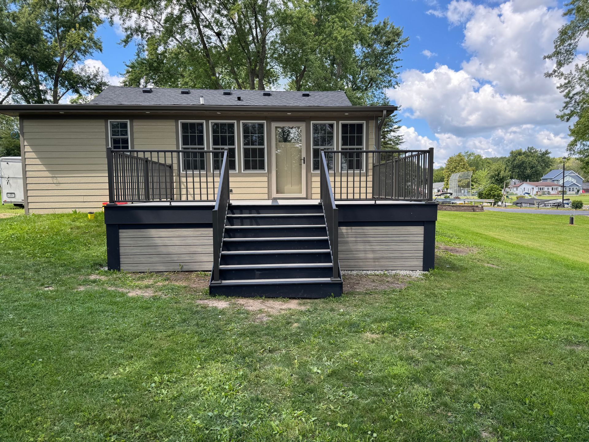 Beige house with a black deck and stairs on a grassy lawn under a partly cloudy sky.