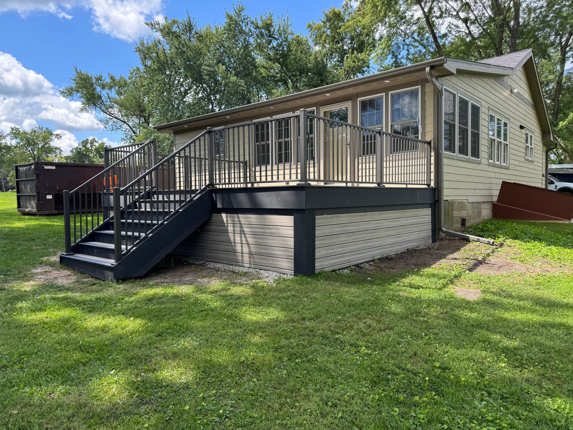 House with gray deck and stairs, white siding, and tan brick base, set on a grassy lawn under a partly cloudy sky.