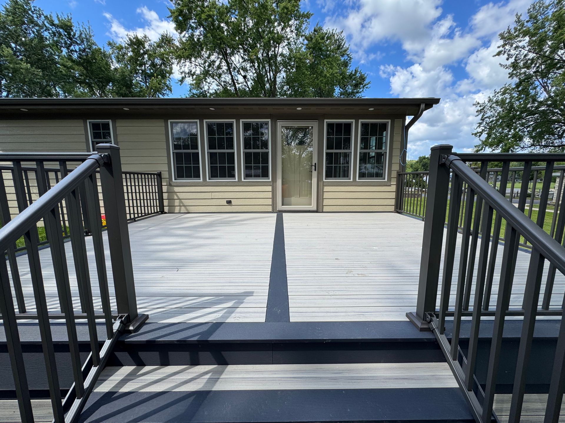 A gray deck with dark railings leads to a light-colored house with windows and a door, trees in the background.