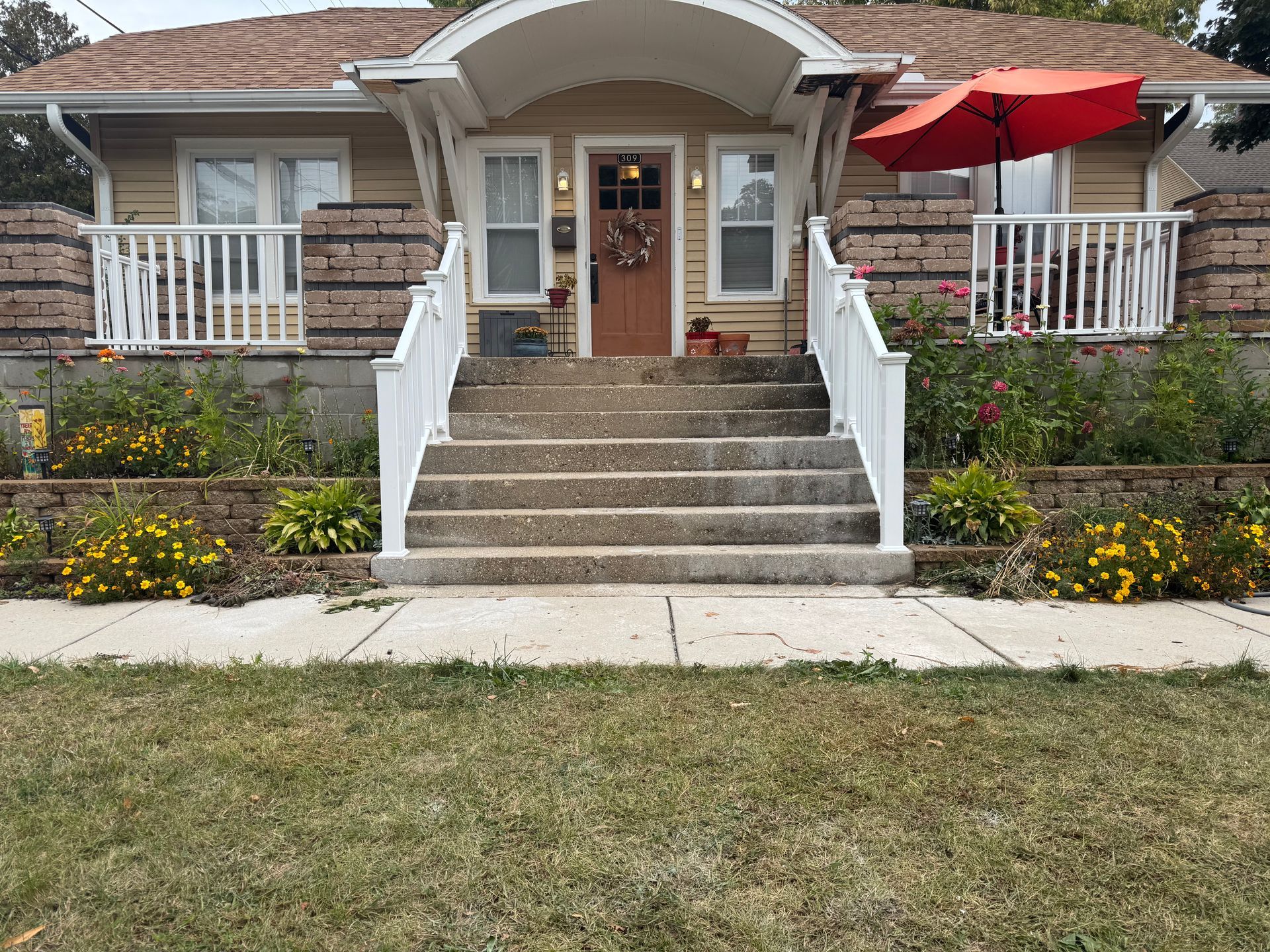 Tan house with steps leading up to the front door, flanked by flower beds and white railing. Orange umbrella on porch.
