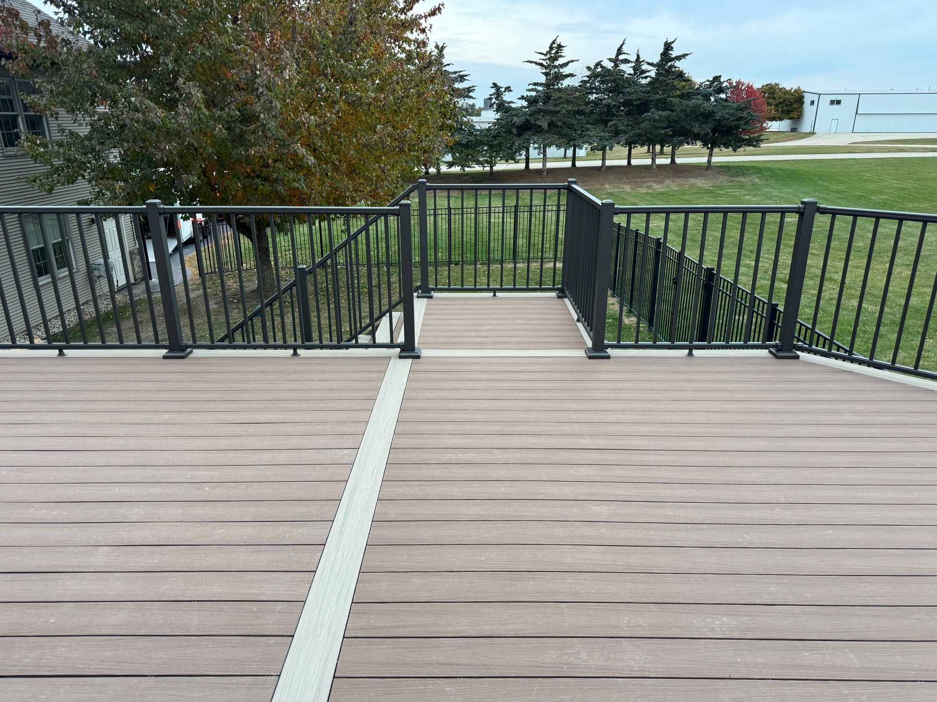 A brown composite deck with black railing overlooking a green lawn with trees and a building against a cloudy sky.