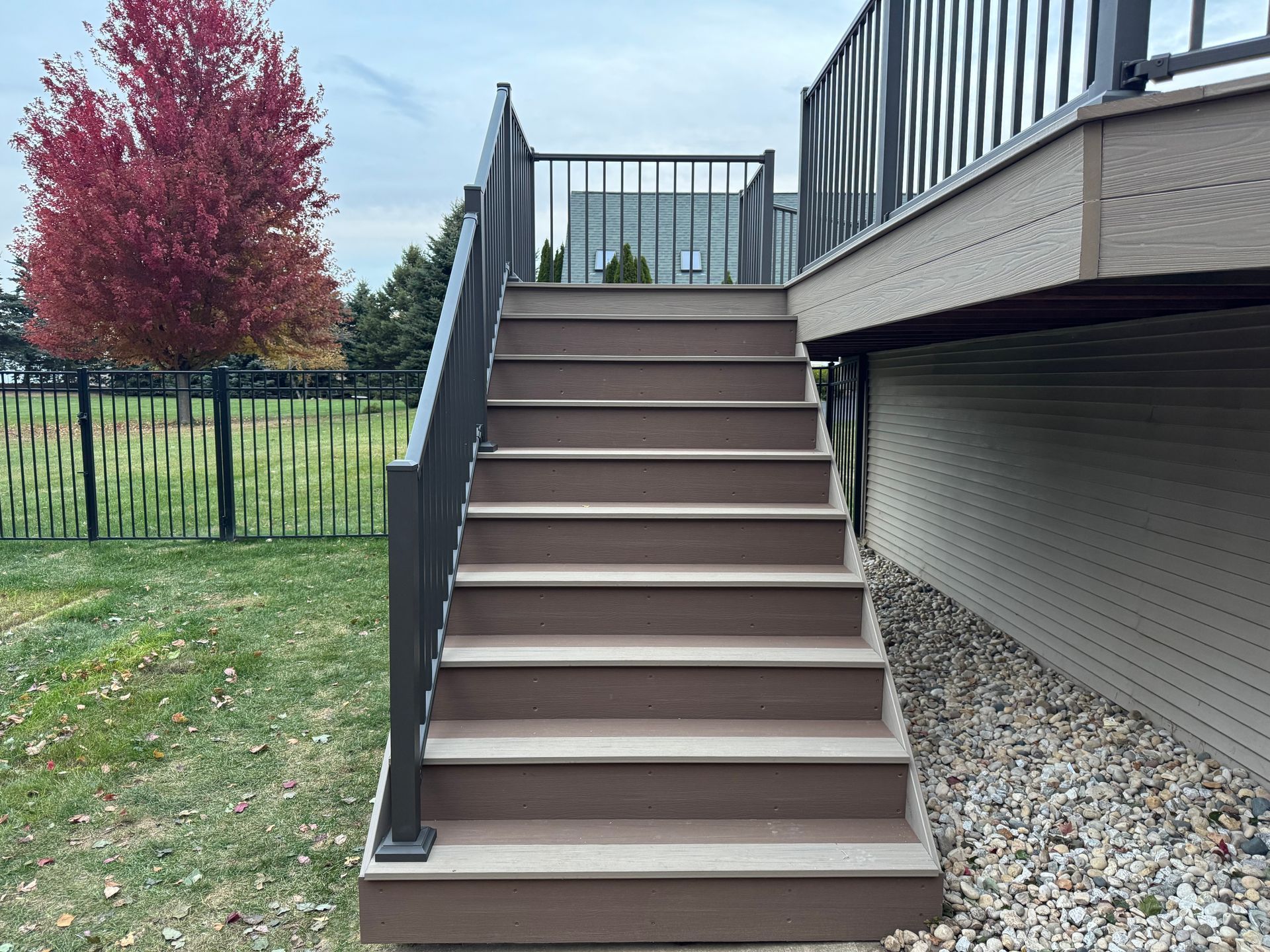 Staircase with brown composite steps leads up to a deck, black railing, beside a grassy yard and a colorful tree.