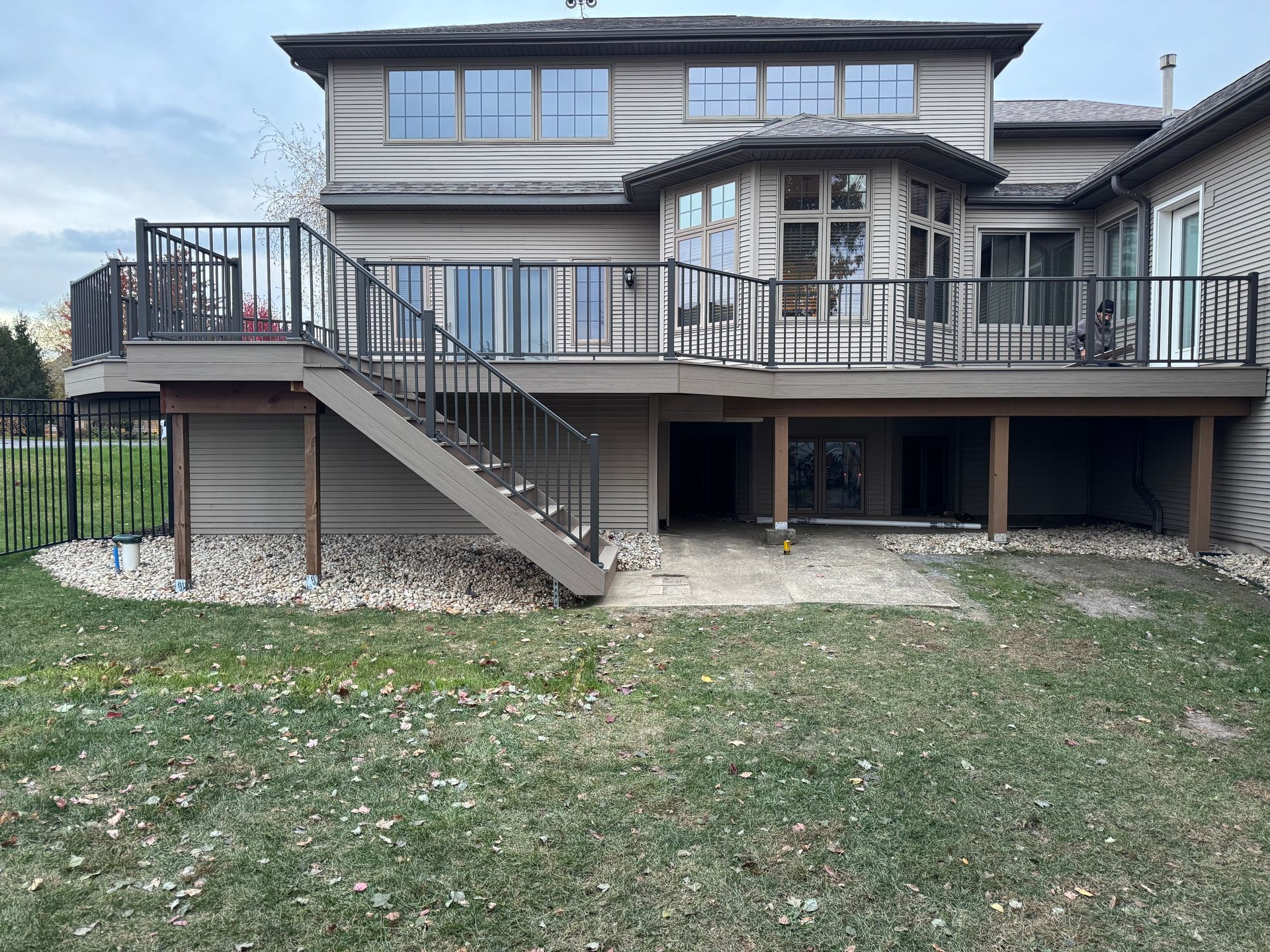 Back view of a two-story beige house with a wooden deck and staircase. Gravel and grass surround the house.