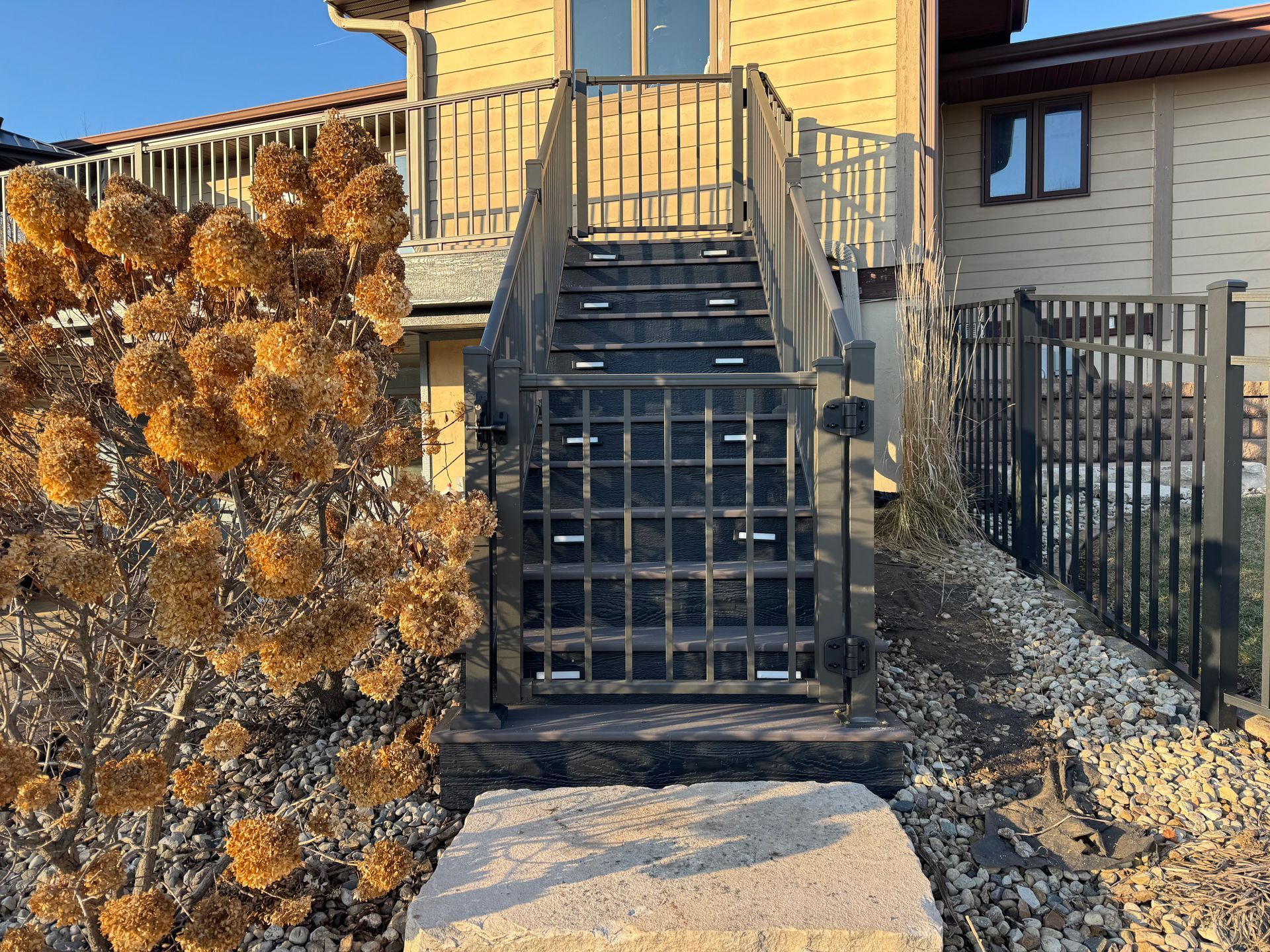 Stone steps lead to a wooden staircase with a gate, in front of a house. Dry bushes on the left.