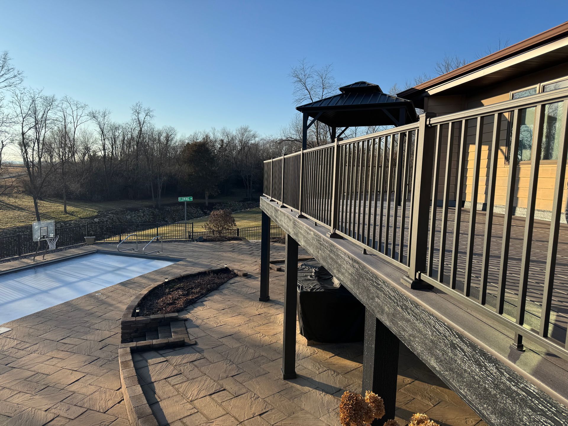Deck with a gazebo, overlooking a patio and a pool covered in a white tarp, trees in the distance.