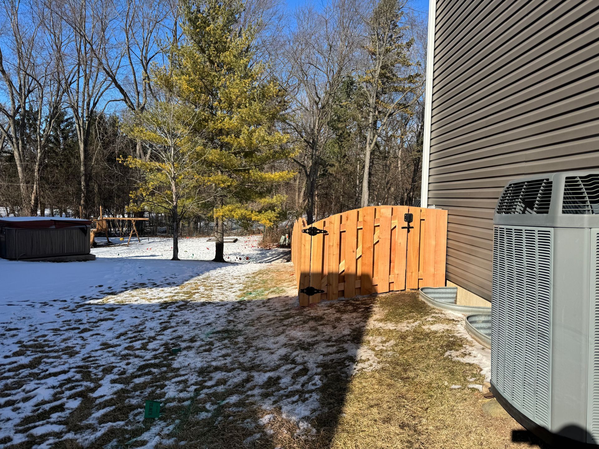 Snowy backyard with a wooden fence, evergreen trees, and an air conditioning unit.