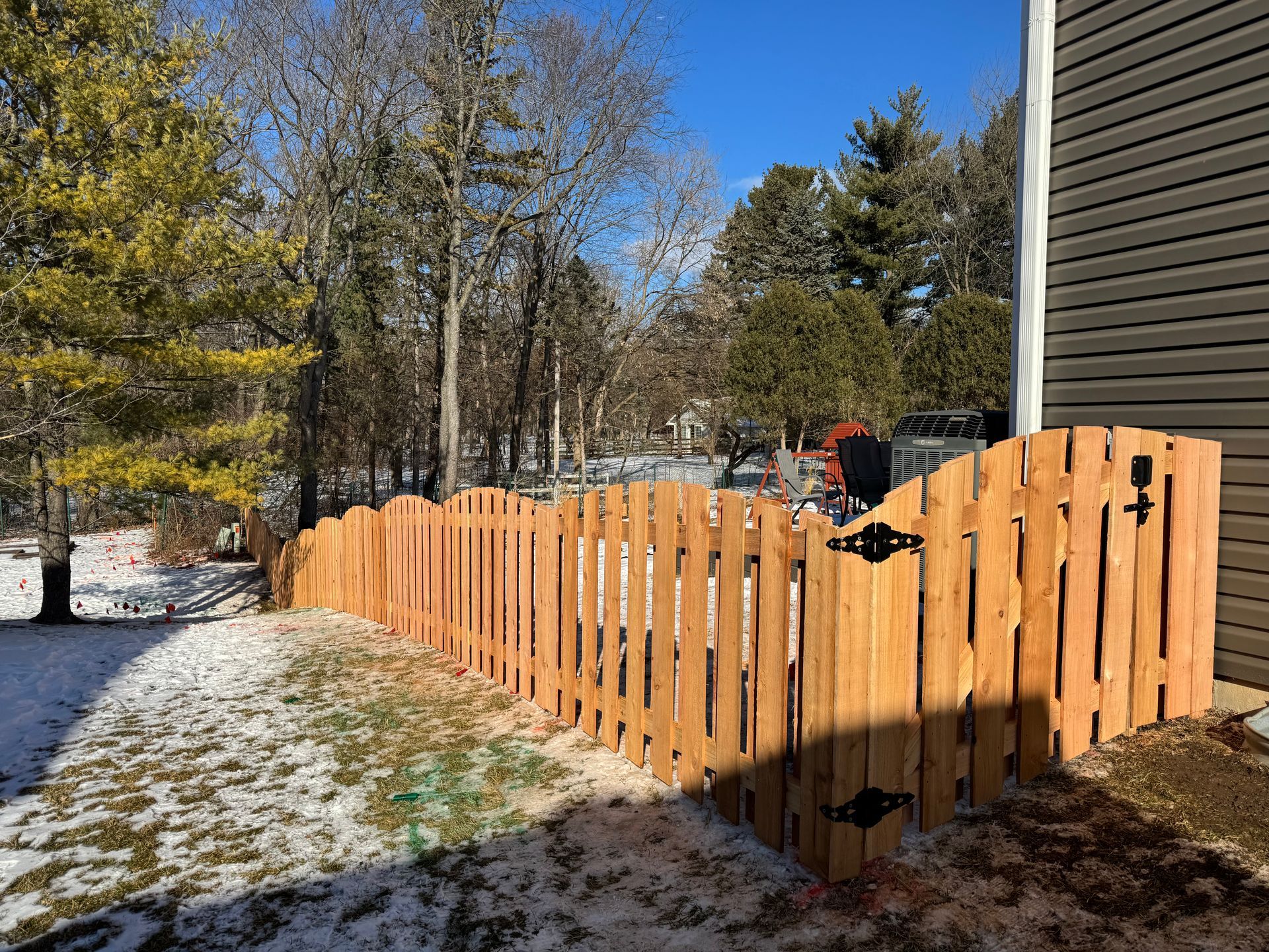 Wooden fence with scalloped top, gate, snow on ground, trees in background, sunny day.