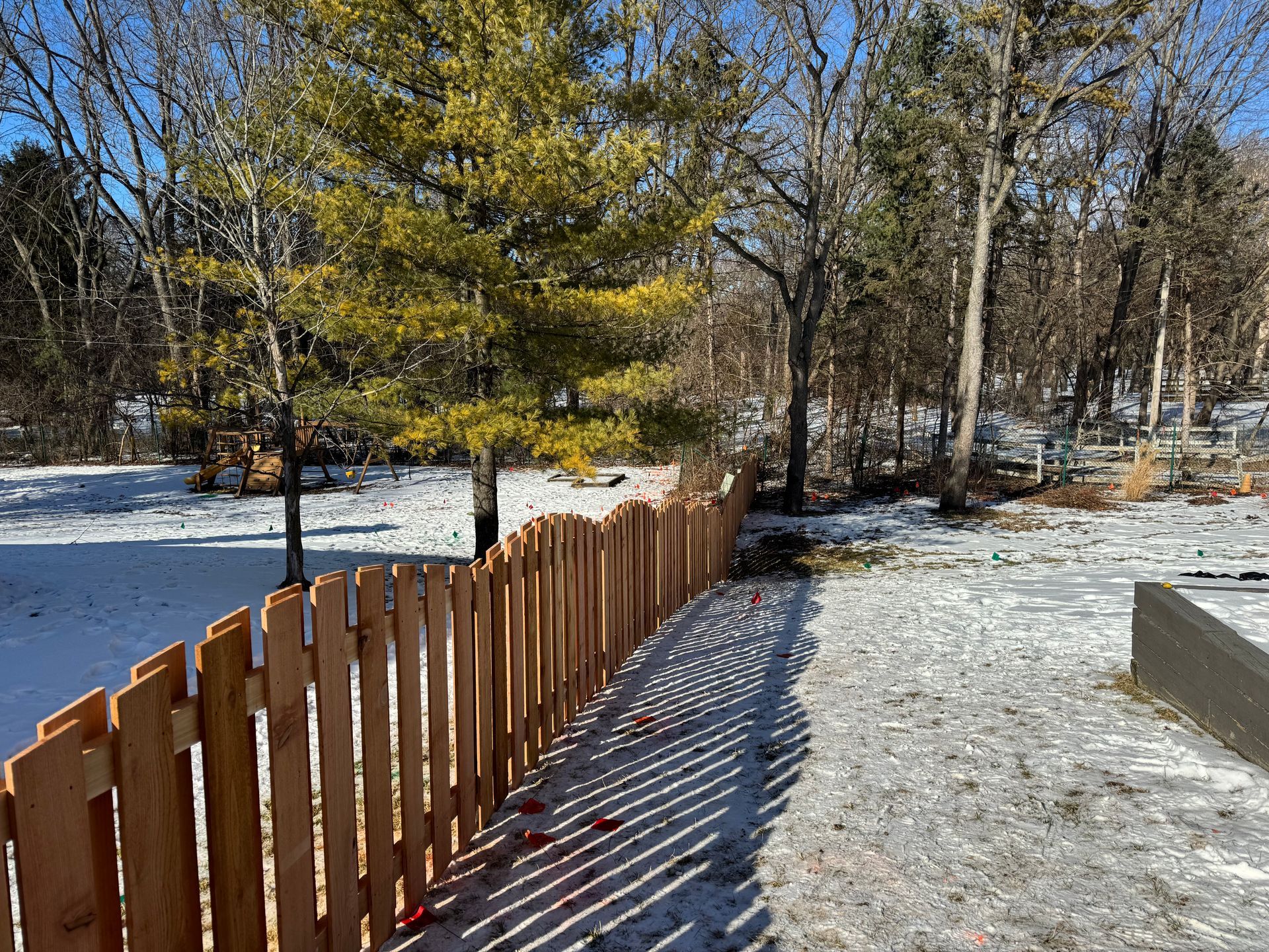 Wooden fence in snow-covered yard, sunny day. Trees in background.