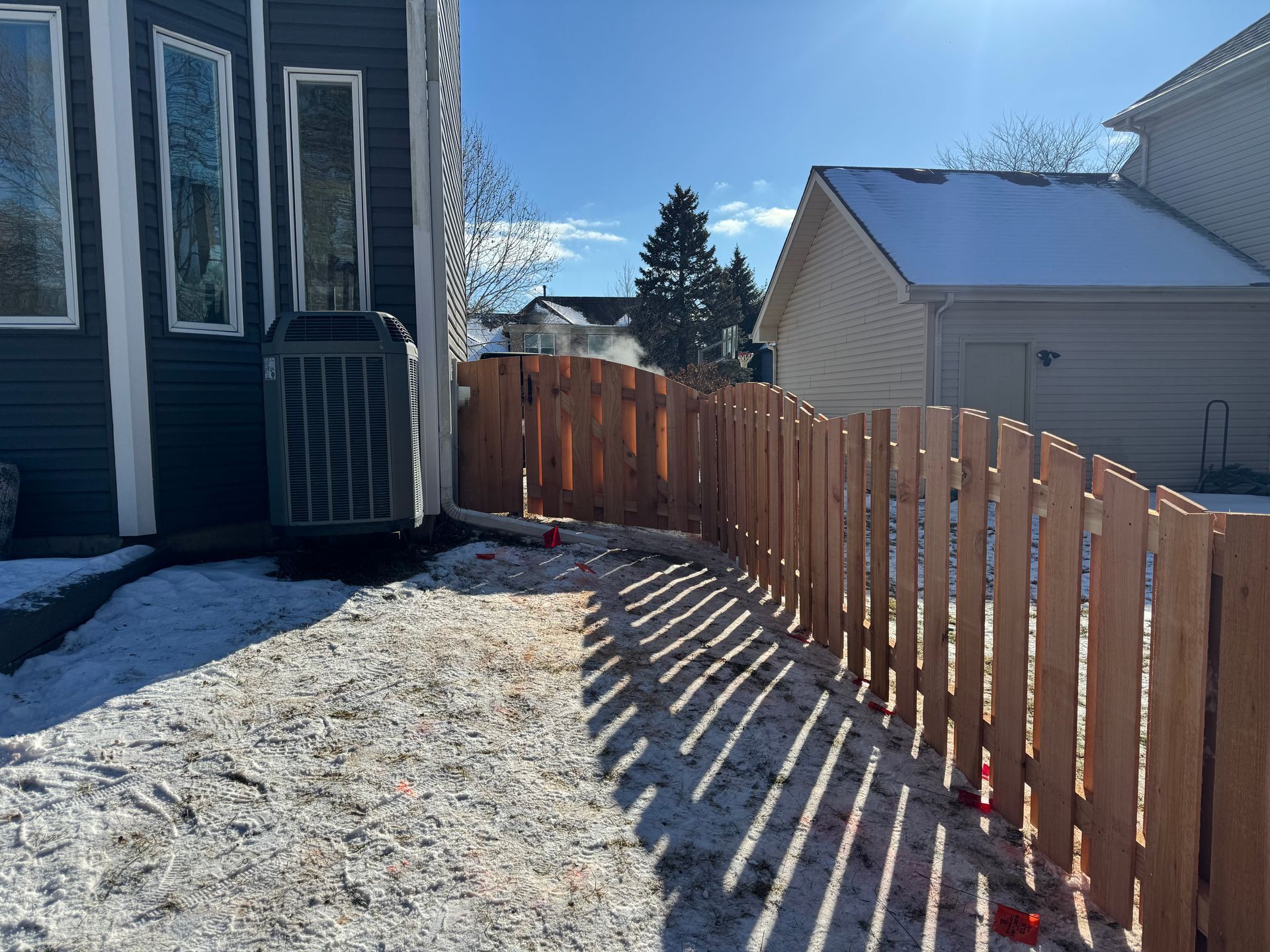 A wooden fence curves along a snowy backyard next to a house with blue siding.