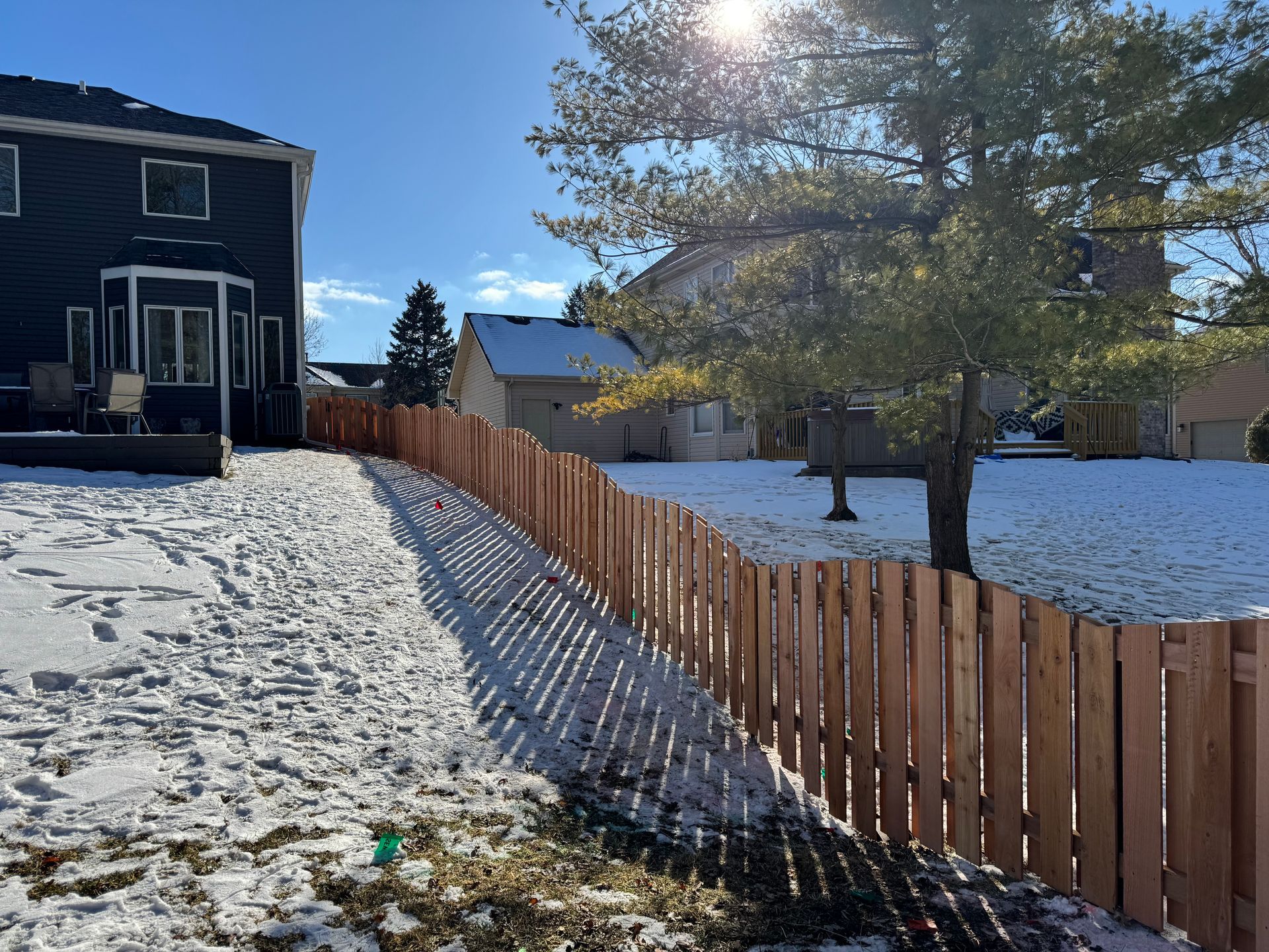 Wooden fence in a snowy backyard with sunlight. Houses and a tree are visible.