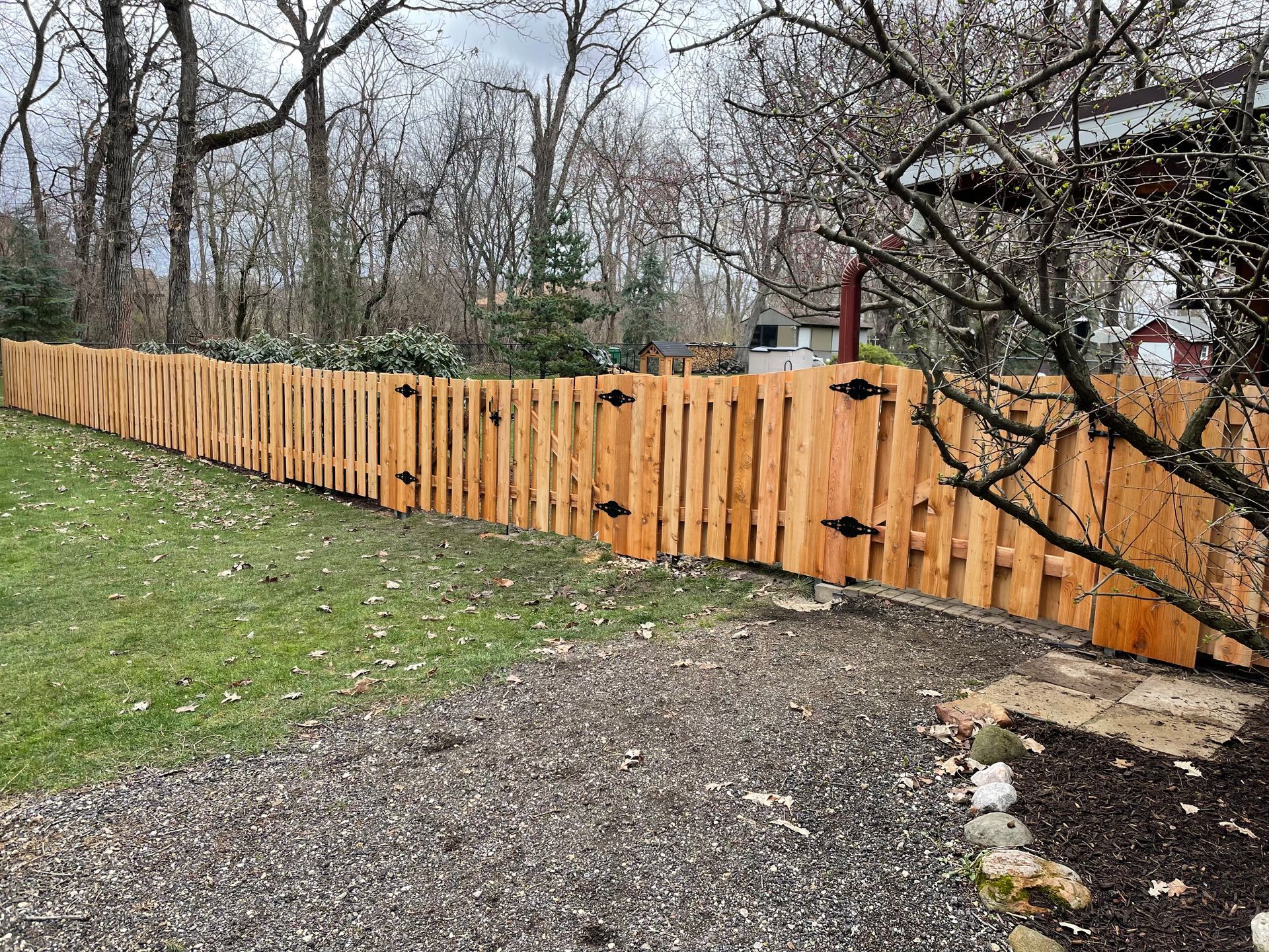 Wooden fence bordering a yard with gravel path and green grass, with trees in the background.