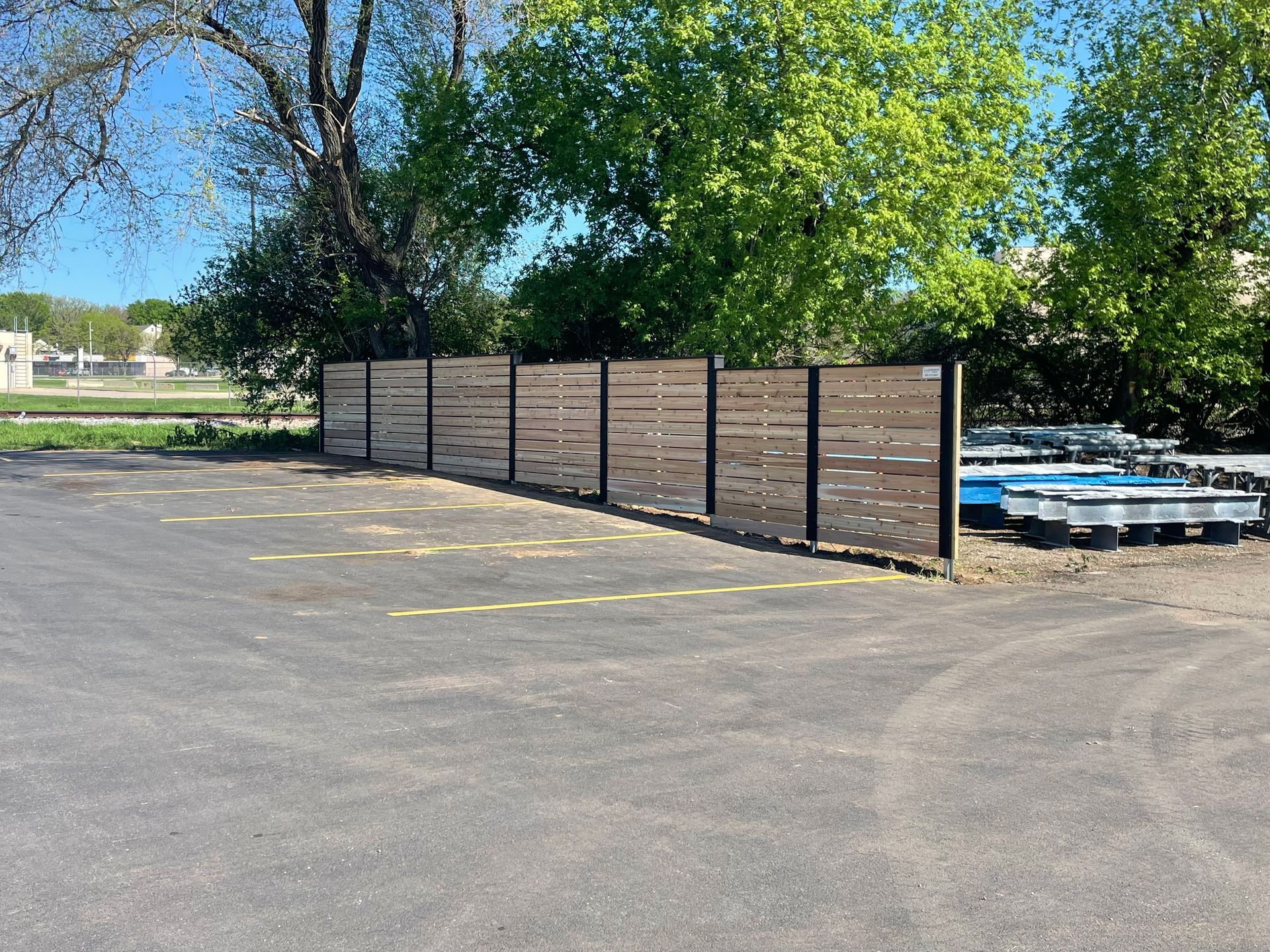 Wooden fence with horizontal slats in a parking lot, trees in the background.