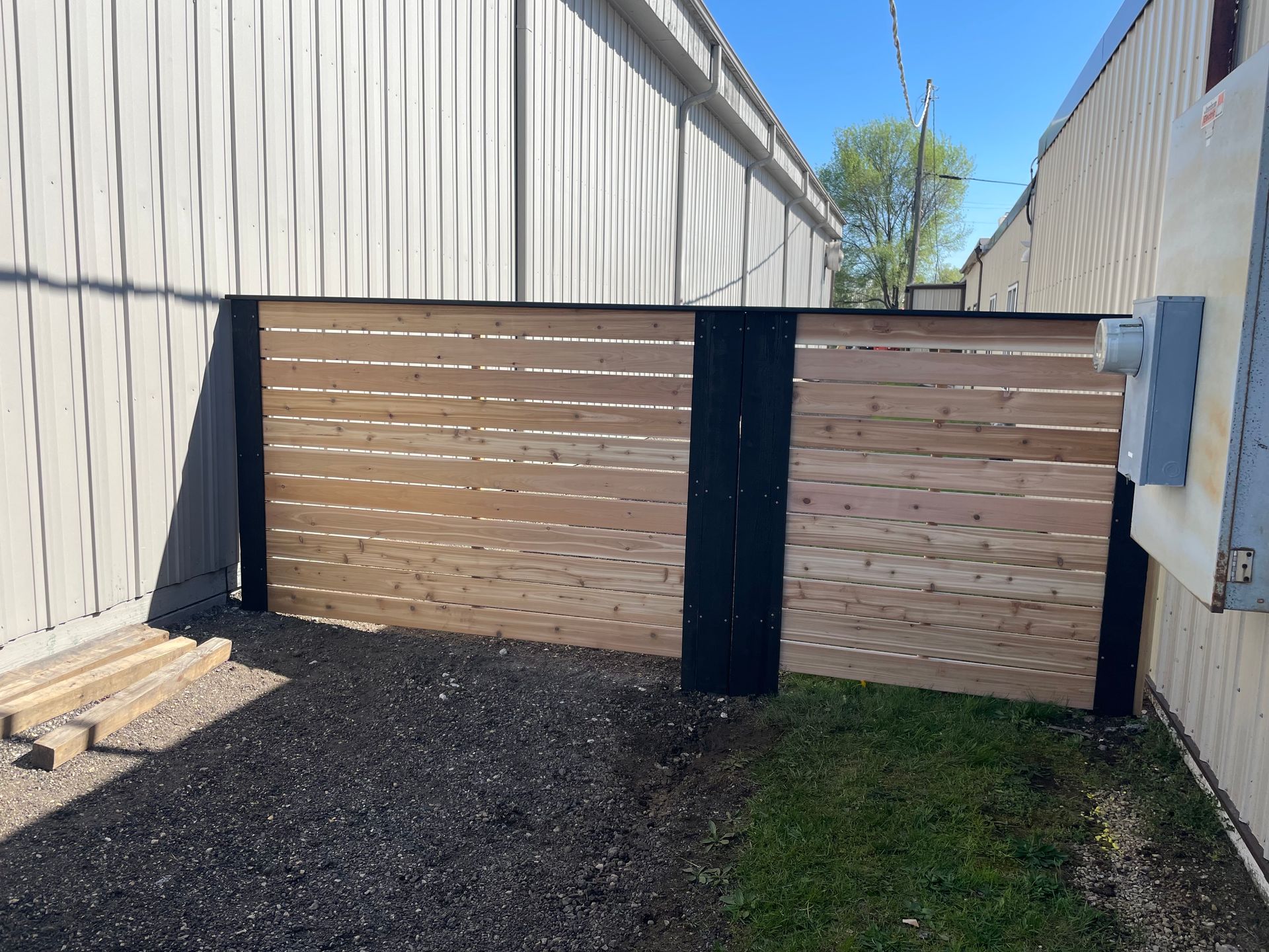 A wooden horizontal slat fence, painted black, separates two buildings on a grassy area.