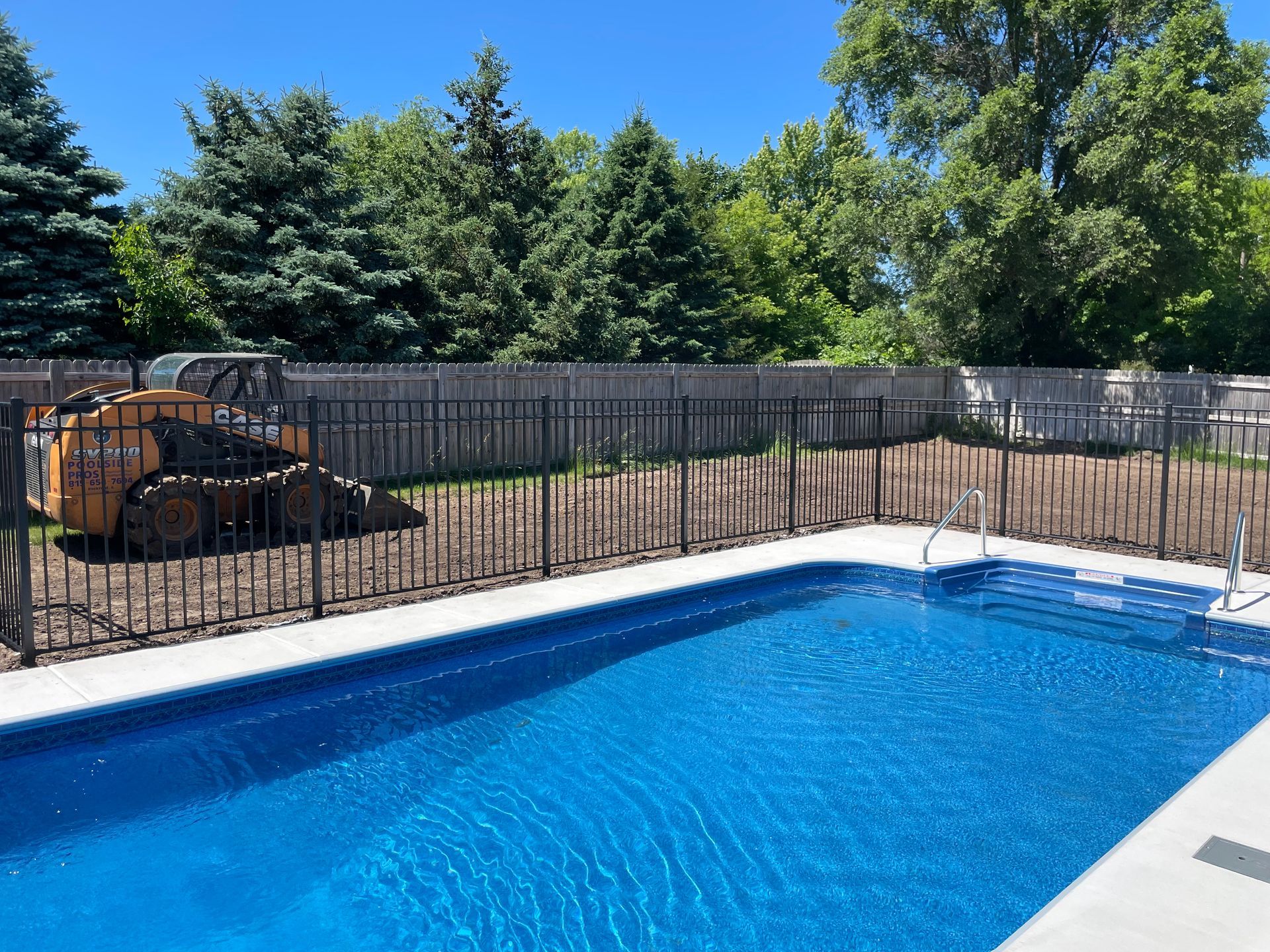 Blue swimming pool with black fence and trees in background; small earthmover on the left. Sunny day.