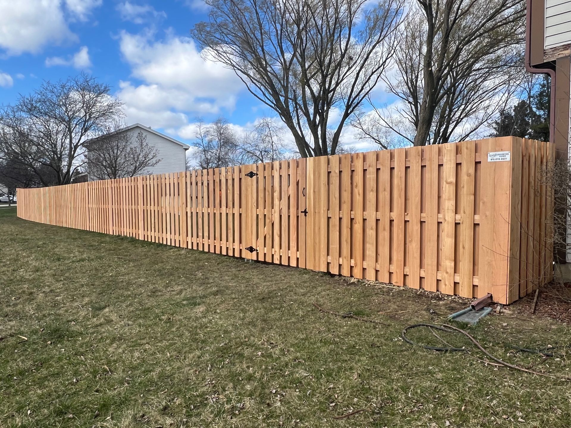 Wooden fence in a grassy yard under a partly cloudy blue sky. Trees and a building are visible in the background.