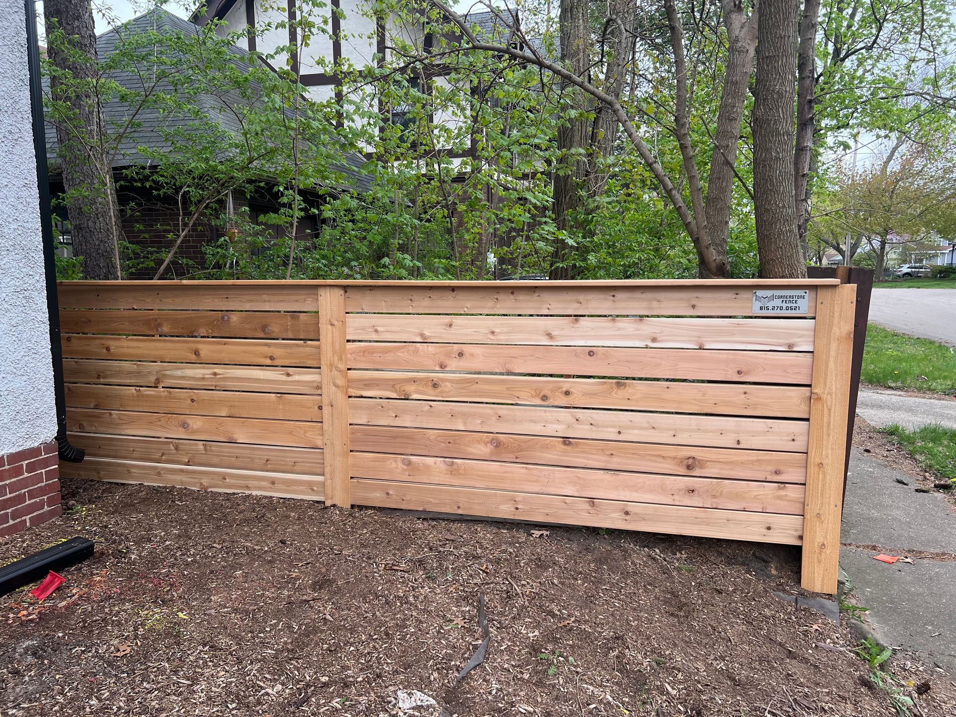 Wooden fence along a sidewalk, separating a yard from the street. Brown wood, horizontal slats. Trees in background.