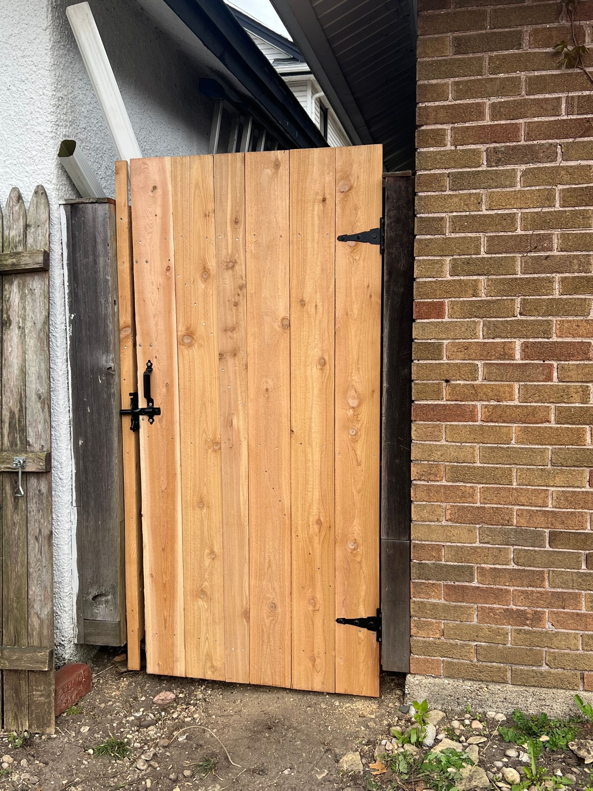 Wooden gate with black hinges and latch, between a brick wall and a fence.