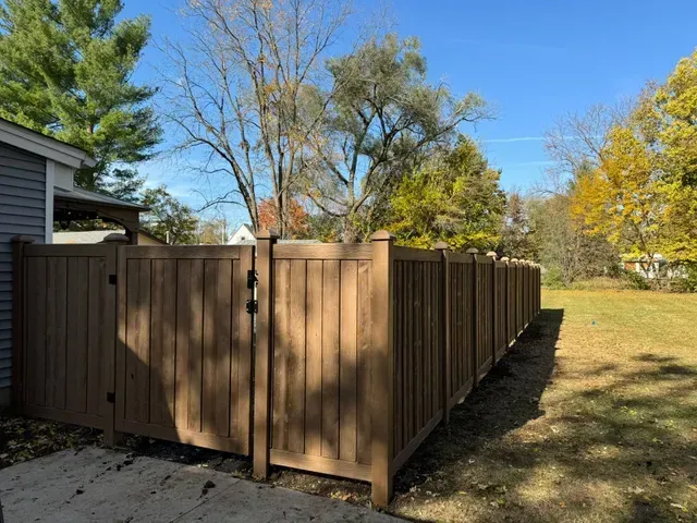 Brown wooden fence with gate in front of a house and yard on a sunny day.