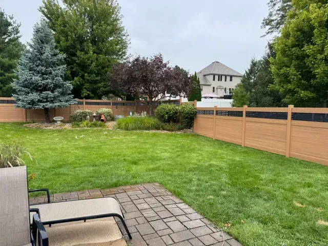 Backyard with brown and black fence, green grass, patio, and trees under a cloudy sky.