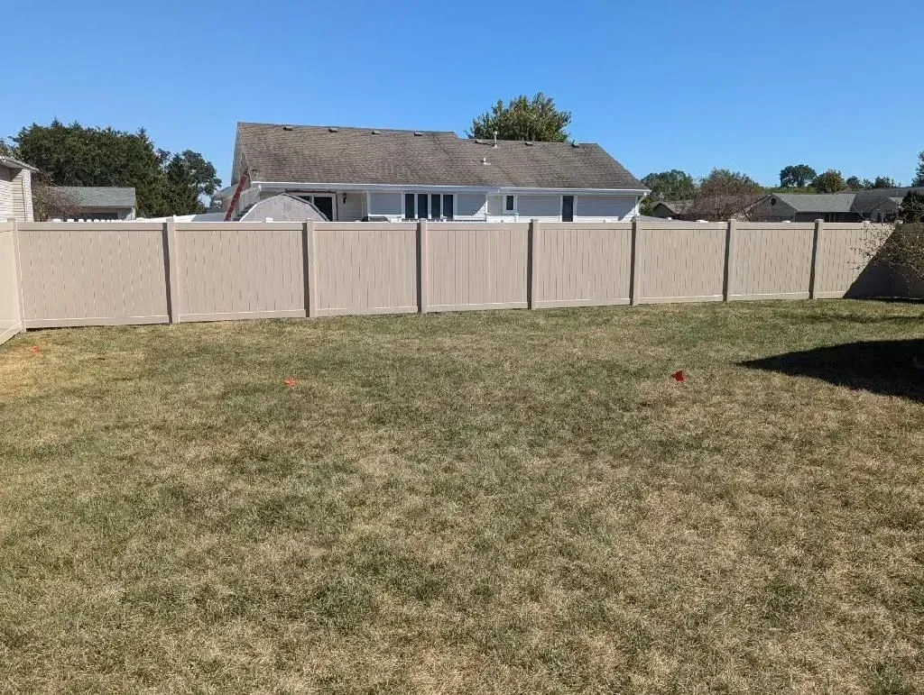 Tan privacy fence surrounding a grassy backyard with a house visible in the background under a blue sky.