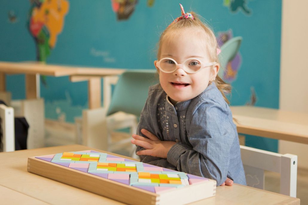 Girl with Down syndrome wearing glasses smiles at a table with puzzle pieces in front of a world map.