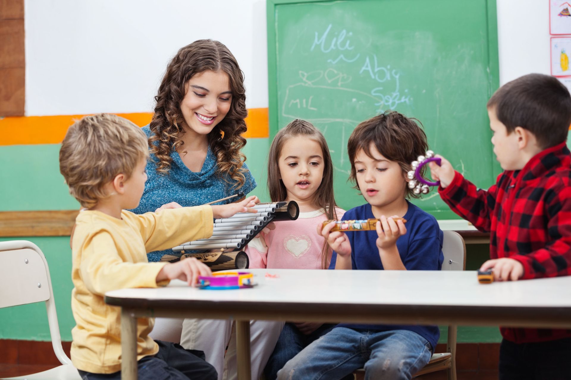 Teacher and children in a classroom playing instruments; xylophone, tambourines. Smiling teacher, focus on music.
