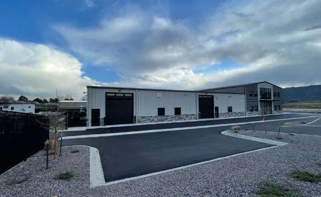 A large white building with black doors is sitting next to a gravel road.