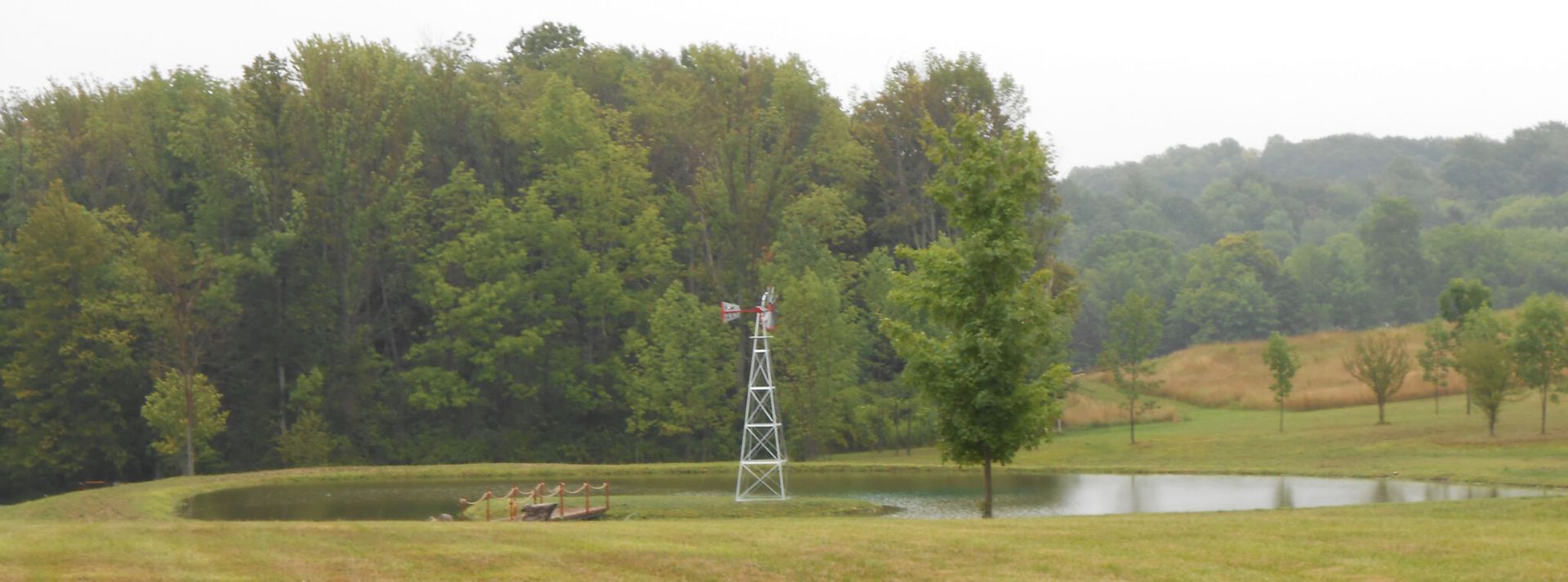 A pond in a field with a windmill, trees, and rolling hills in the background. Overcast sky.
