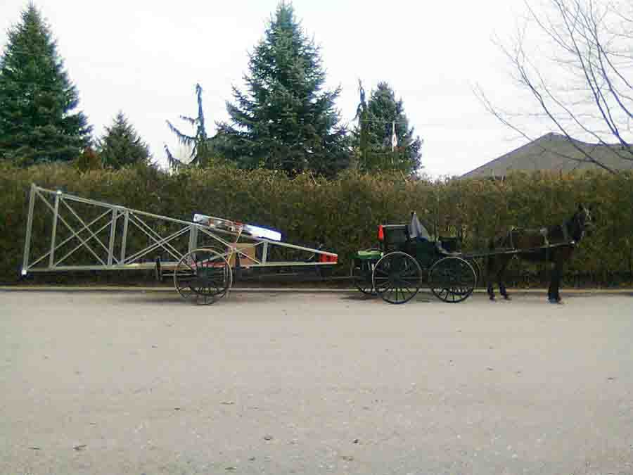 Horse-drawn carriage pulling a trailer with a tall, lattice-like structure outdoors on a paved road.