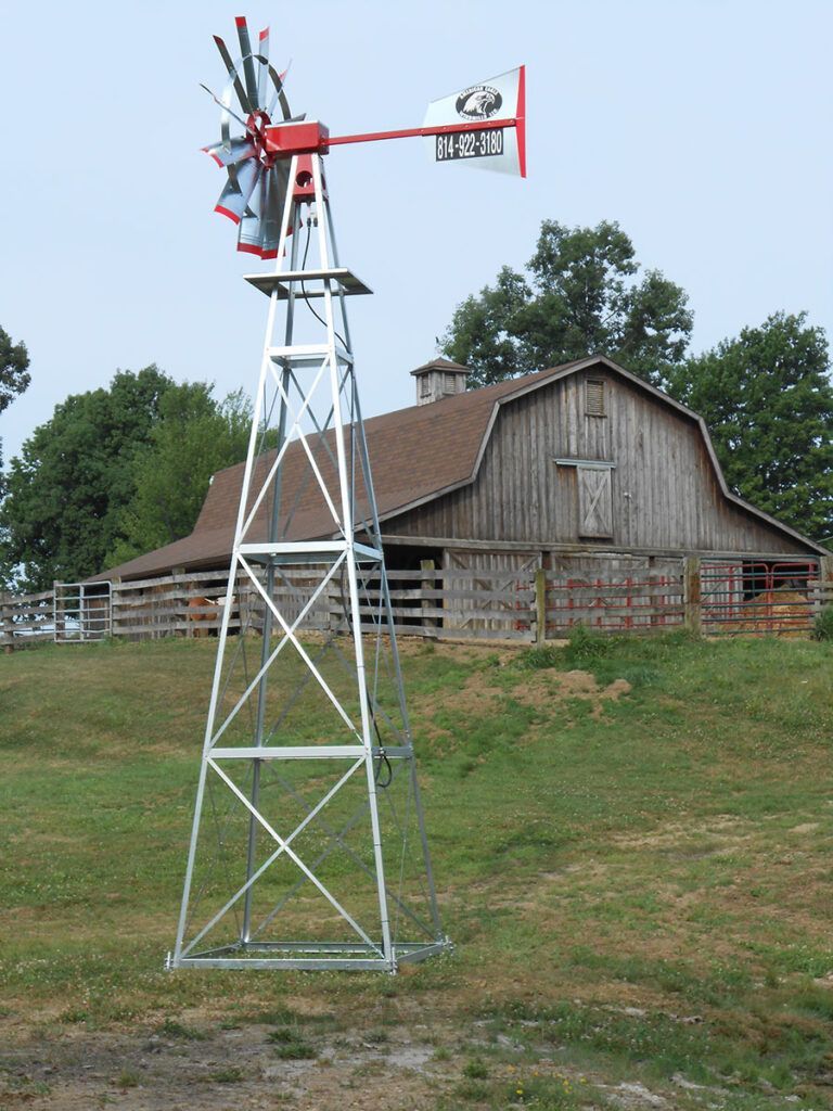 Windmill on a metal tower, with a red top, and a barn in the background on a grassy hill.