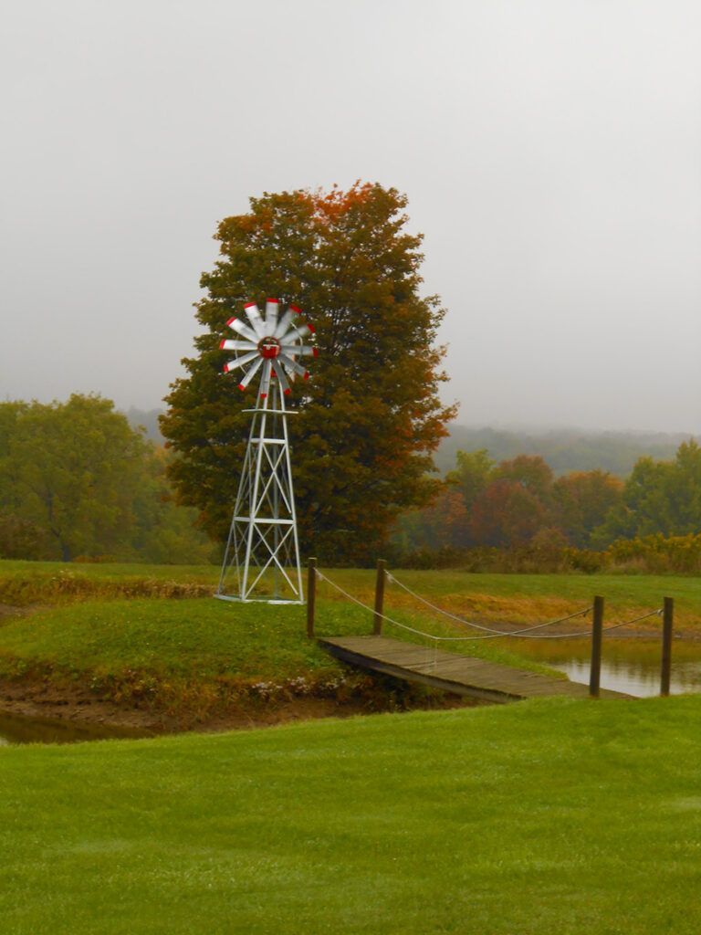 Windmill on a green lawn by a small bridge and pond, with colorful fall foliage. Cloudy sky.