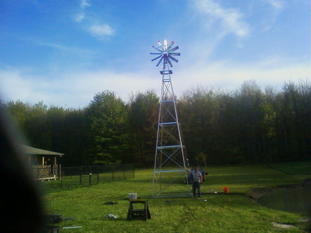 Windmill on a metal tower in a grassy field, trees in the background, a person standing nearby.
