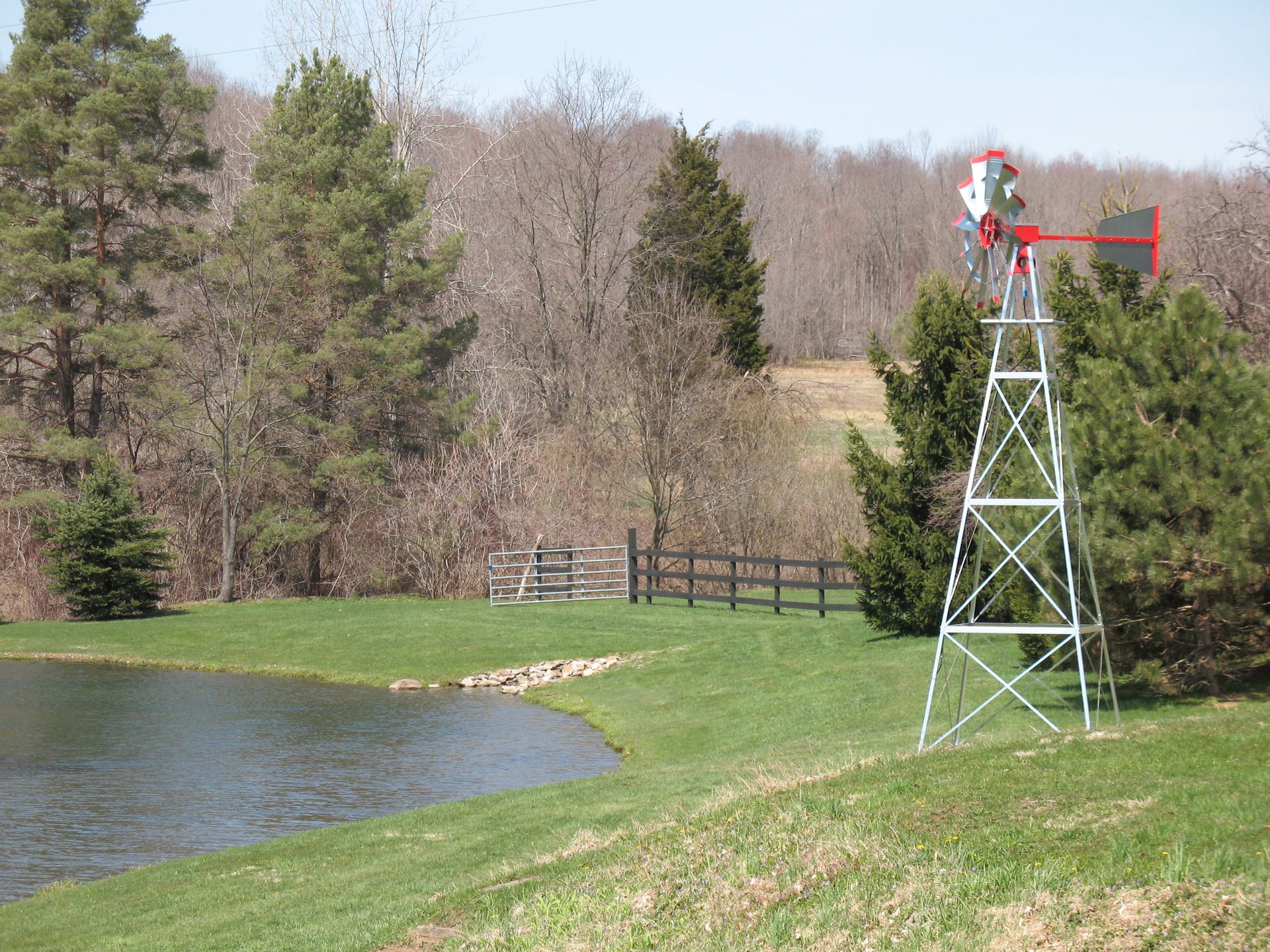 Windmill next to a pond in a grassy yard, with trees in the background under a blue sky.
