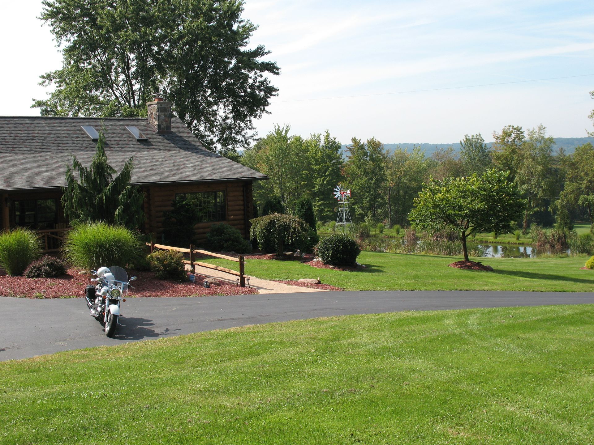 Log cabin with a motorcycle parked in front on a sunny day. Green lawn, trees, and view in the background.