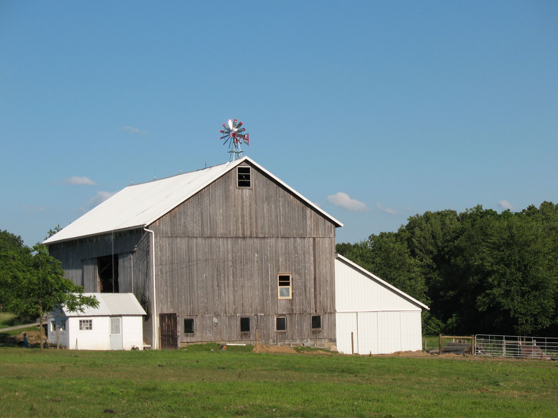 Weathered barn with white roof, small shed, and windmill on a sunny day in a field.