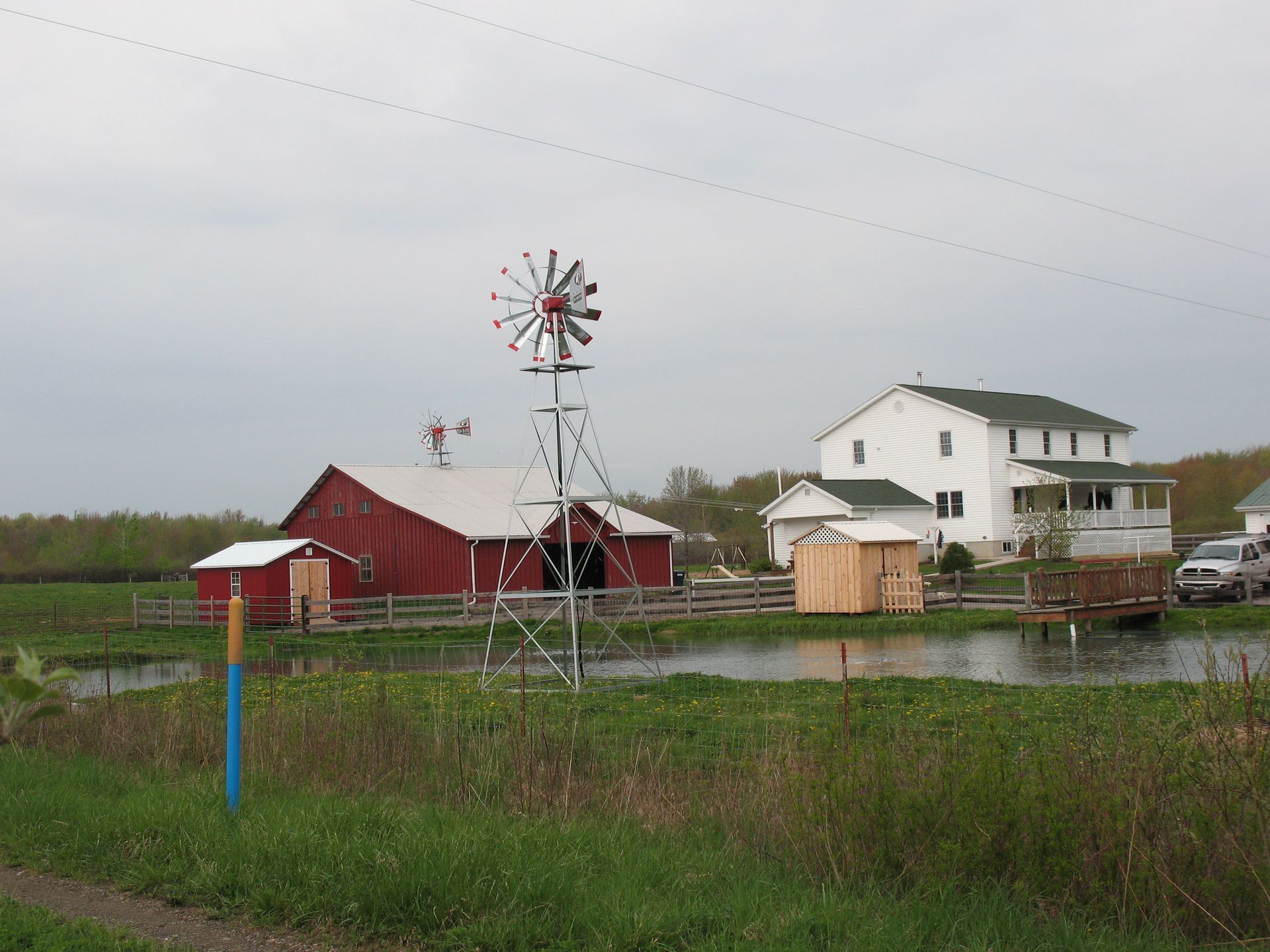Farm scene with red barn, white house, windmill, pond, and overcast sky.
