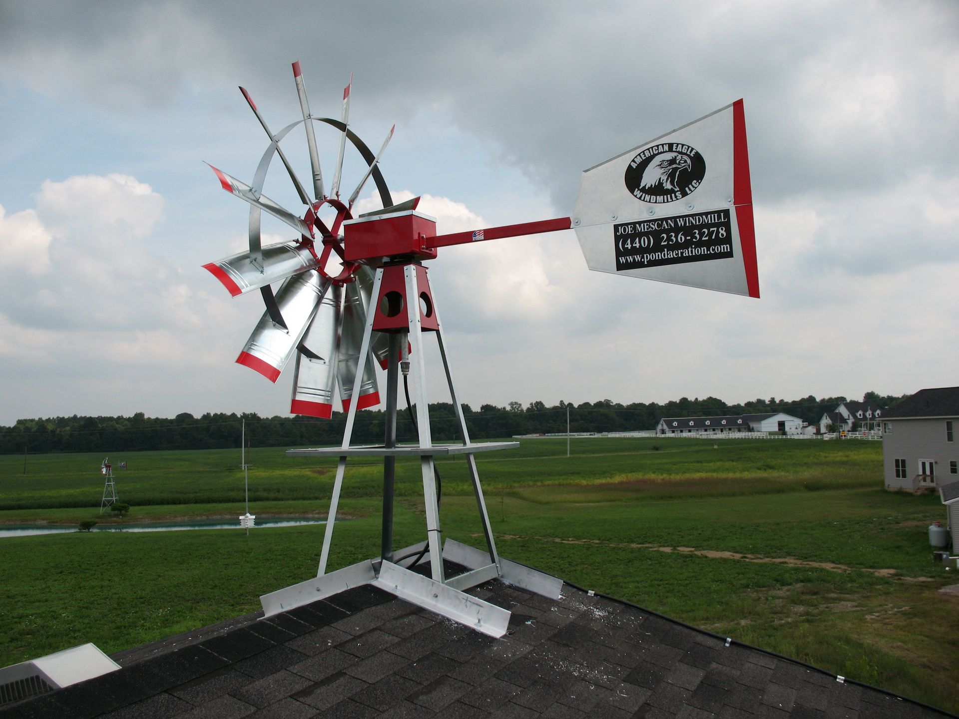 Windmill on a rooftop with red and silver blades and a white and red tail, set against a green field and sky.