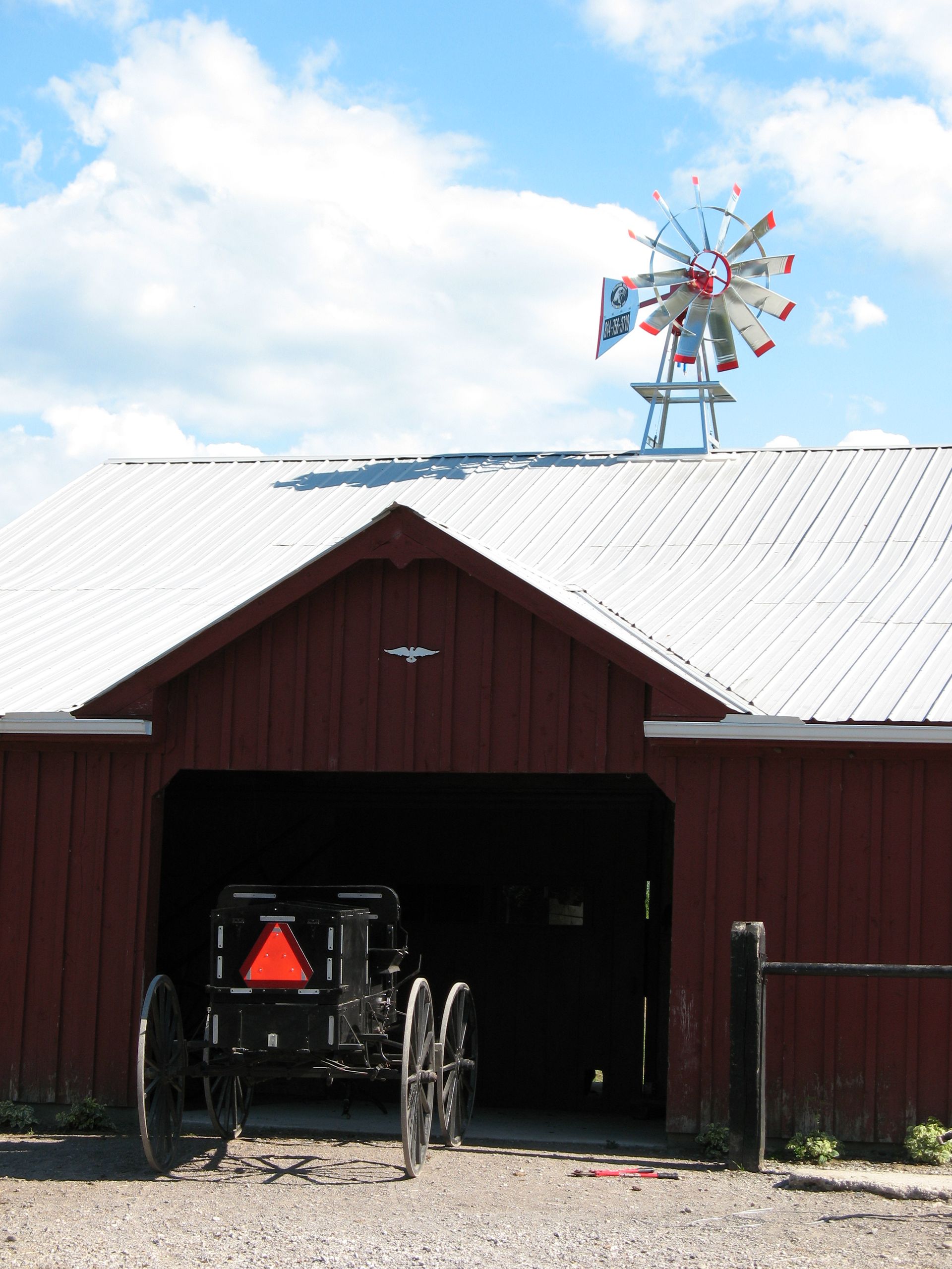 Red barn with black buggy parked inside; windmill on roof, blue sky background.