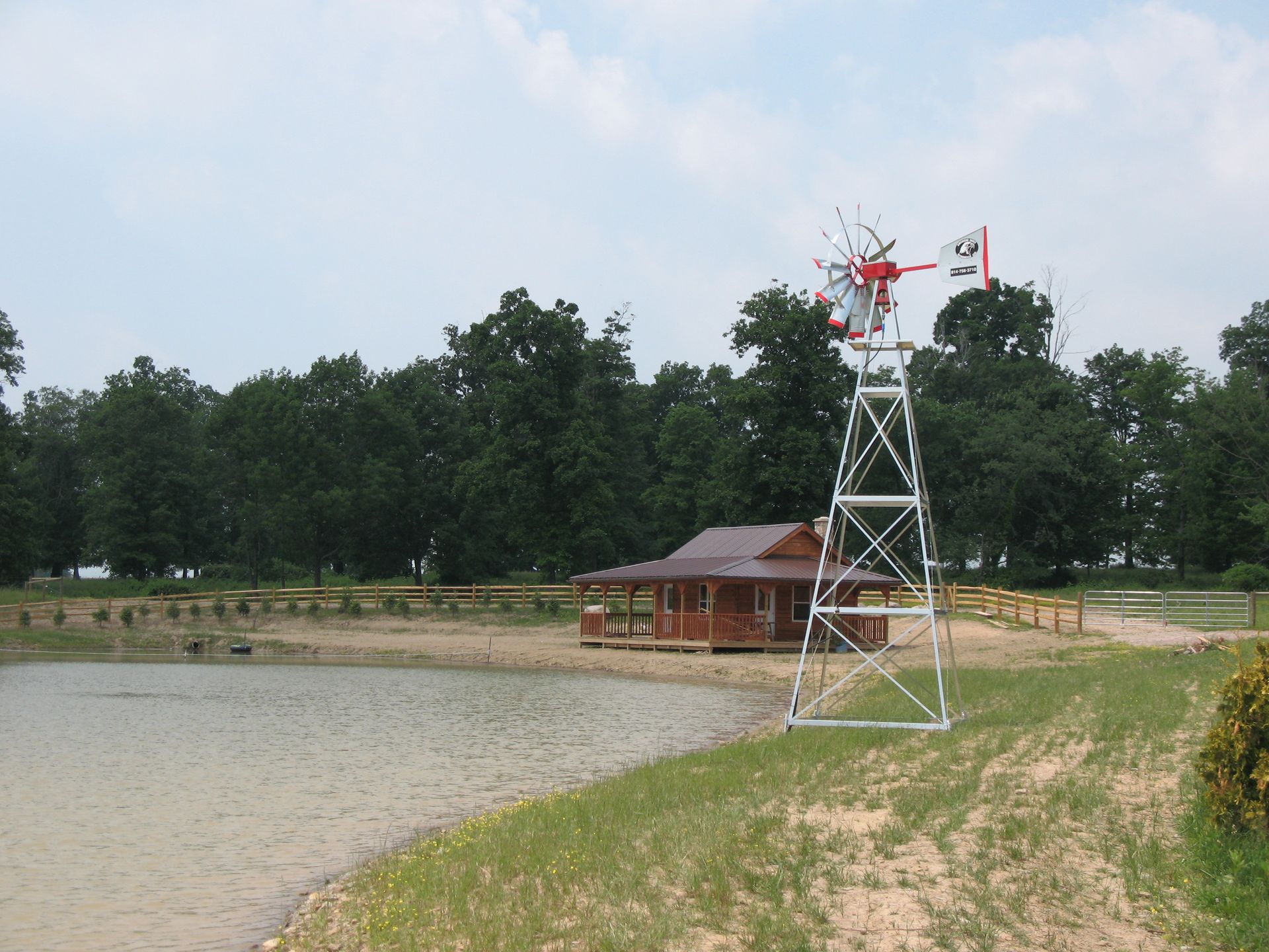 Windmill next to a small wooden building and pond, in front of a treeline under a cloudy sky.