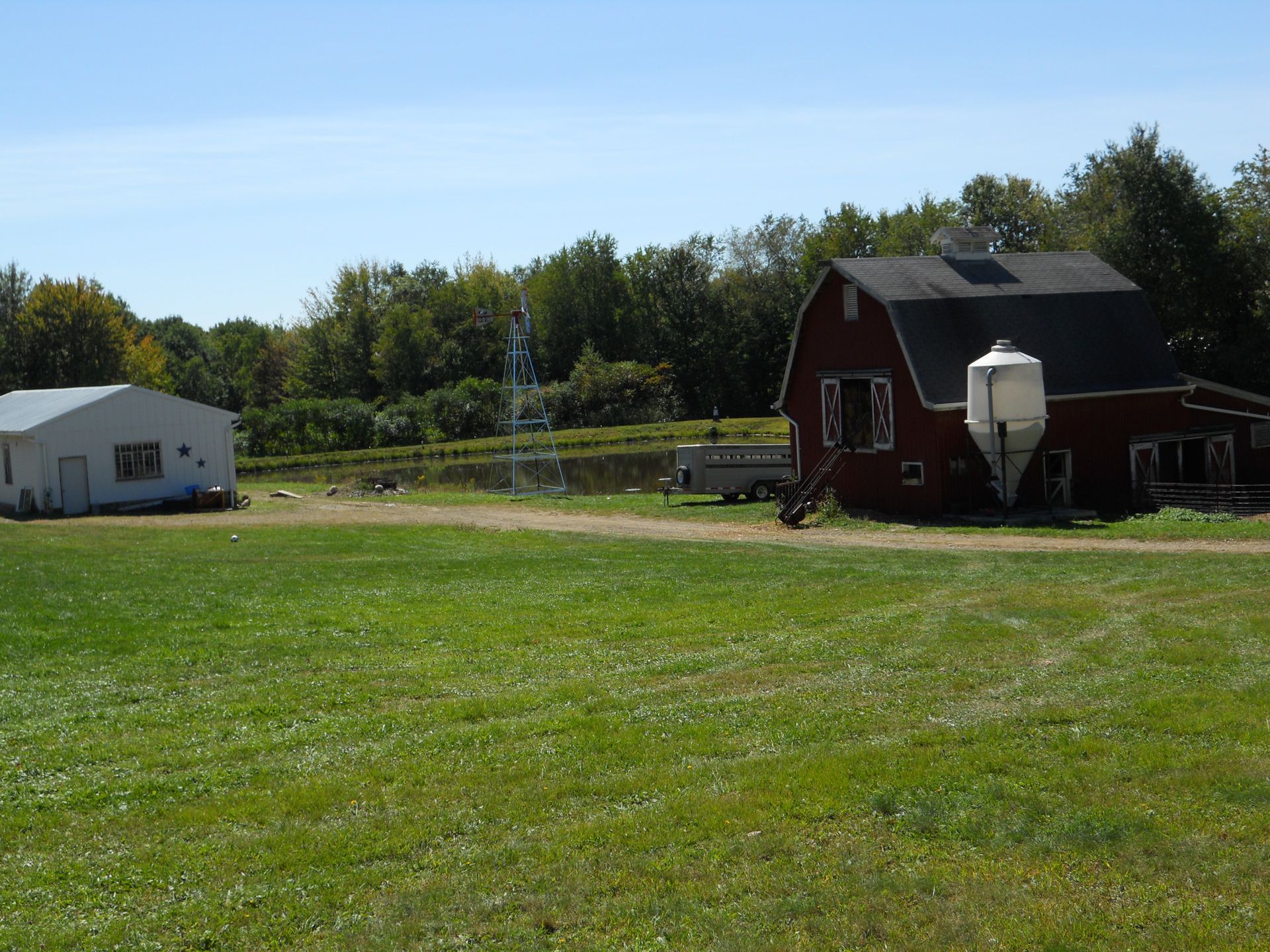 Red barn and white building on a grassy farm, under a blue sky, with trees in the background.